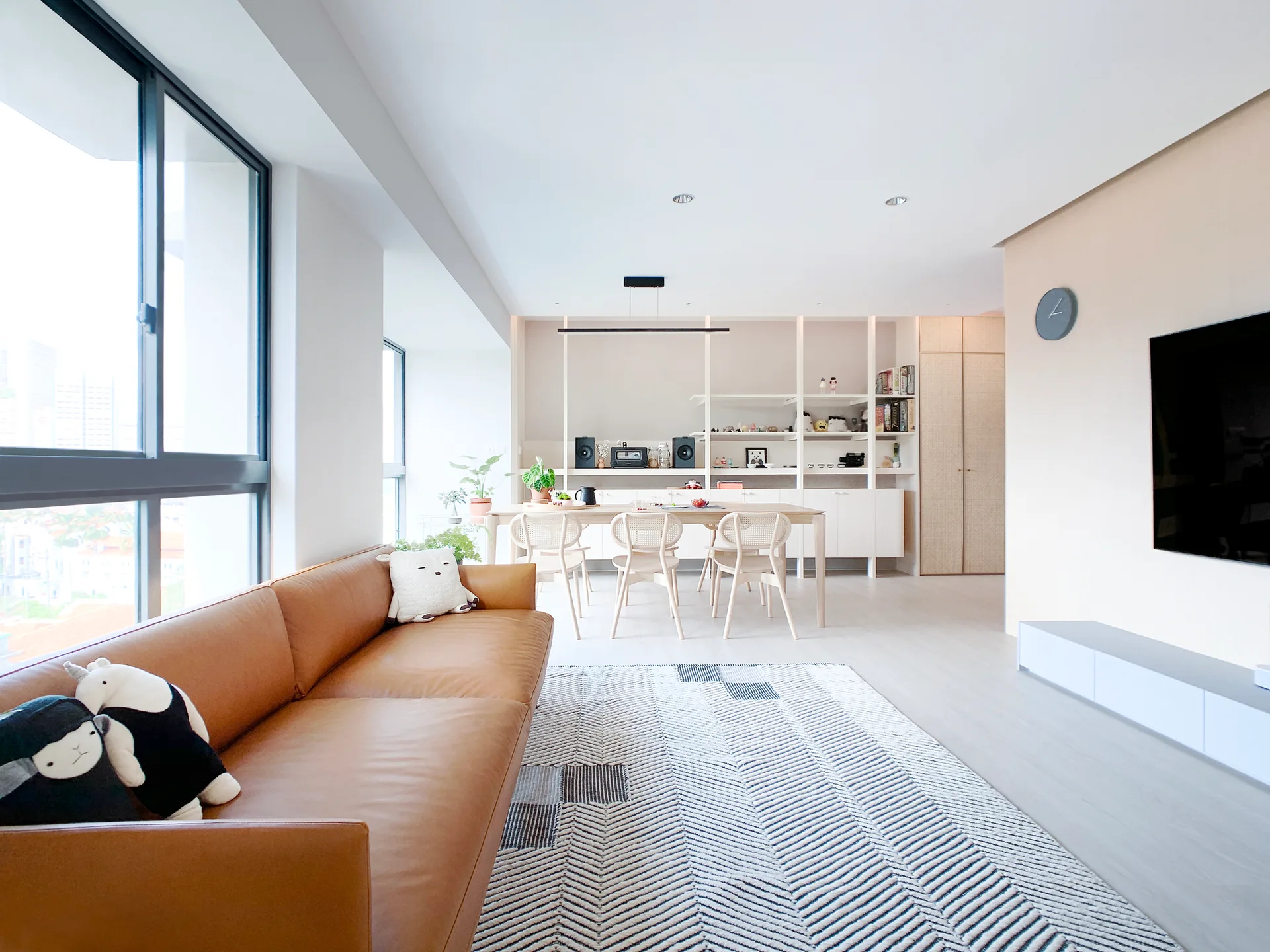 A bright open-plan living room featuring a tan leather sofa, a geometric area rug, and a light wood dining set with built-in shelving.
