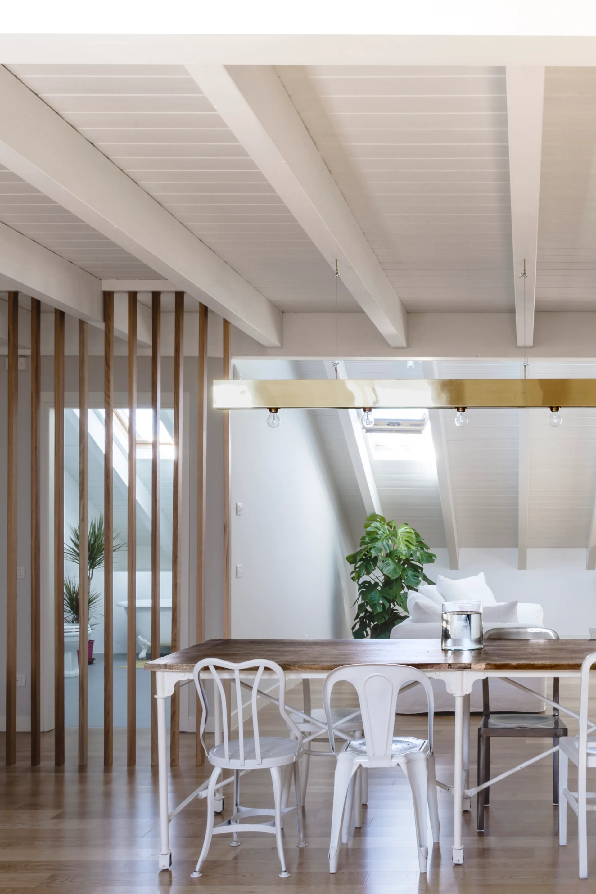 A light-filled dining space features a wood-top table, white metal chairs, a brass pendant light, and a vertical wood slat partition screen.