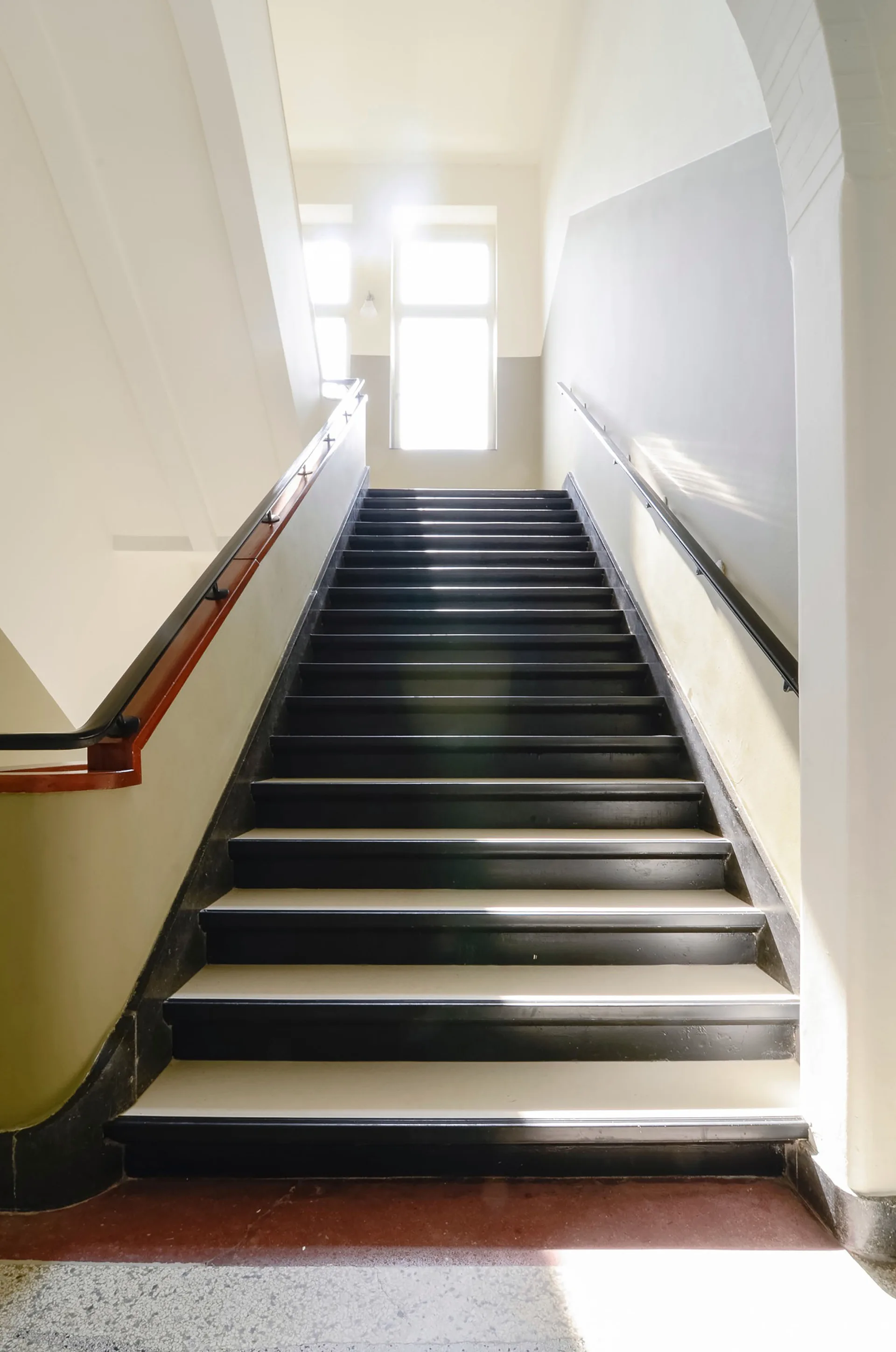 A straight indoor staircase with painted black treads and white risers leads up to a bright window with natural light.