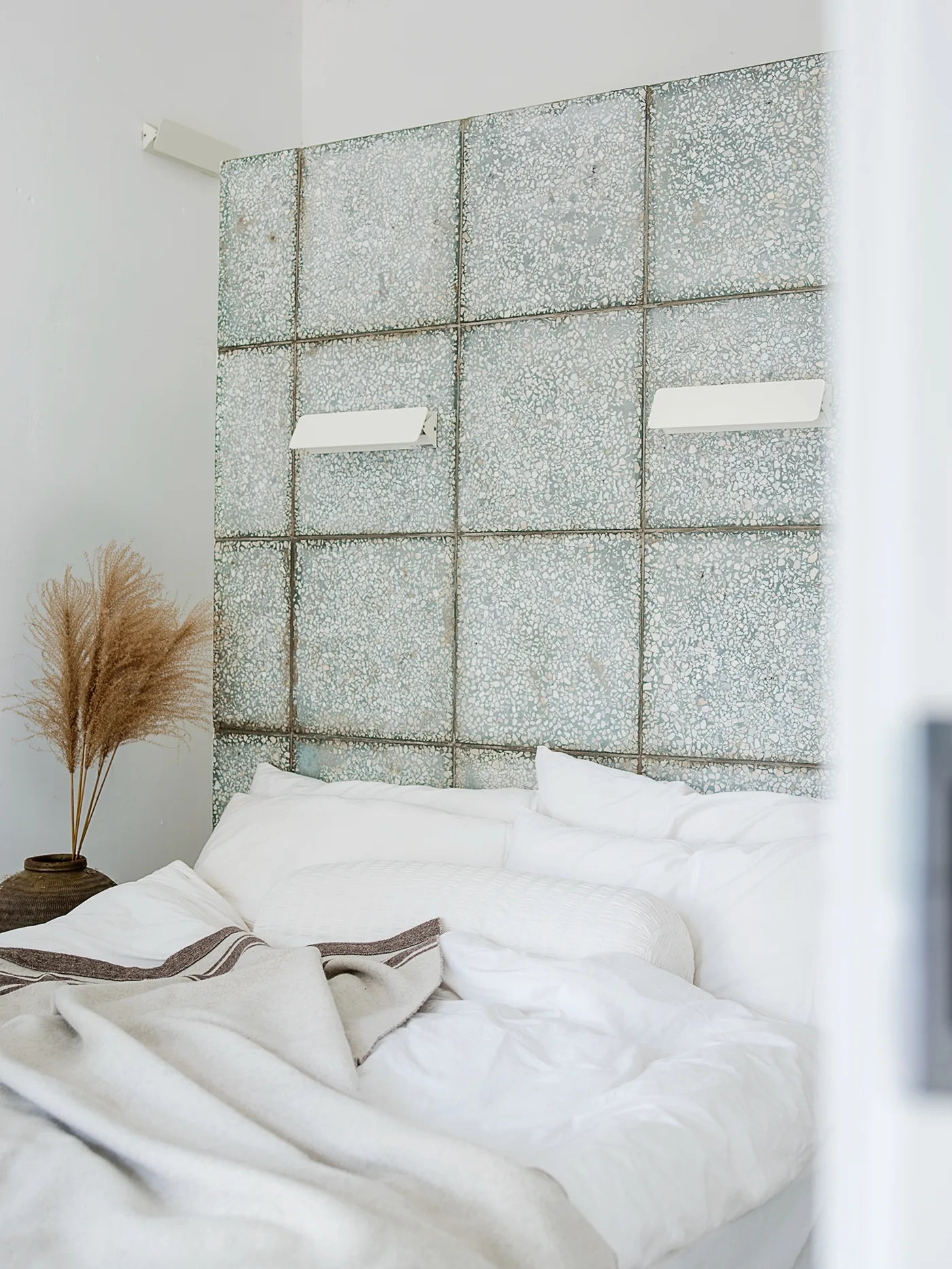 A minimalist bed with white linens sits against a feature wall made of large green terrazzo tiles with two white wall sconces.
