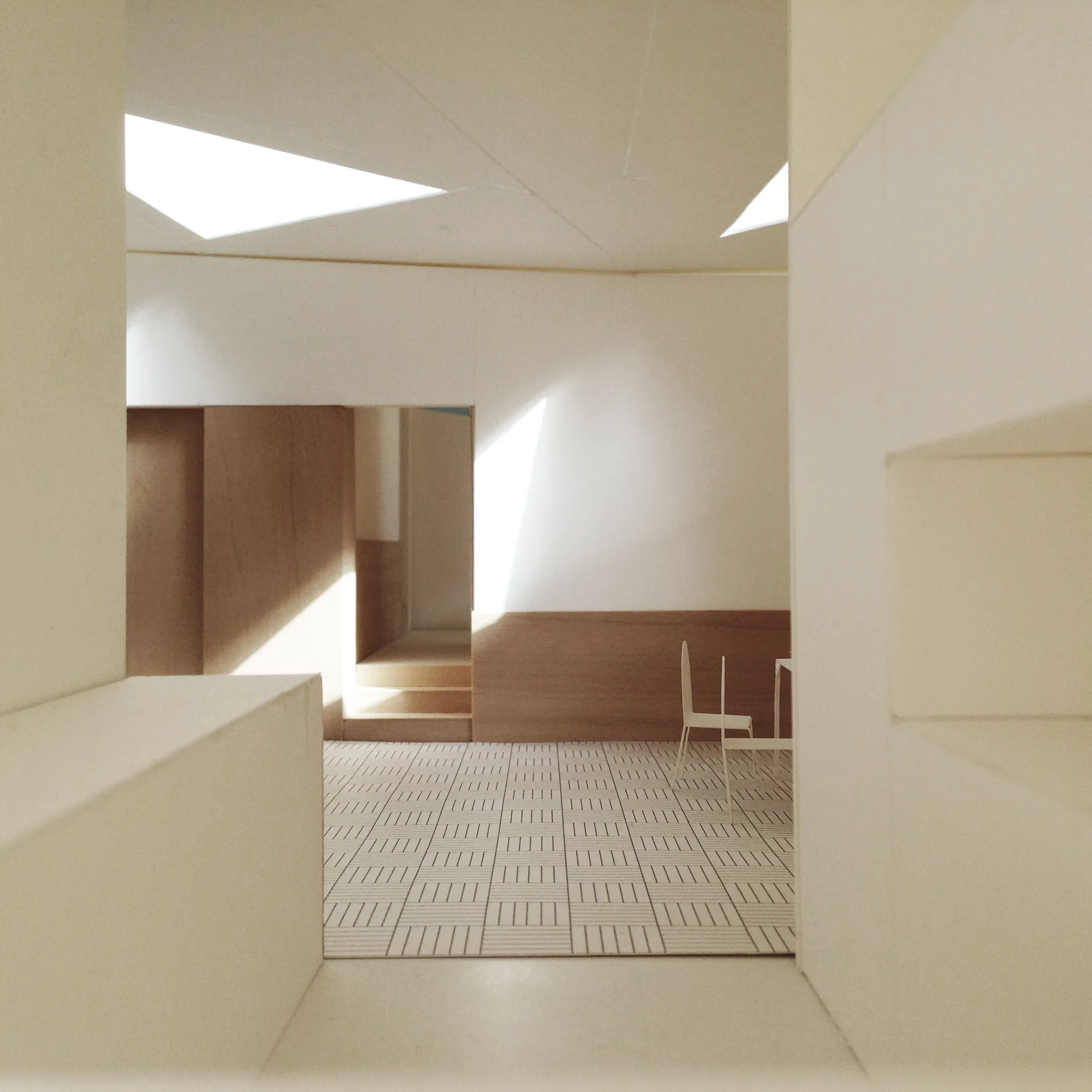 An architectural scale model of a minimalist interior space featuring a grid patterned floor, low wood paneling, and triangular ceiling skylights.