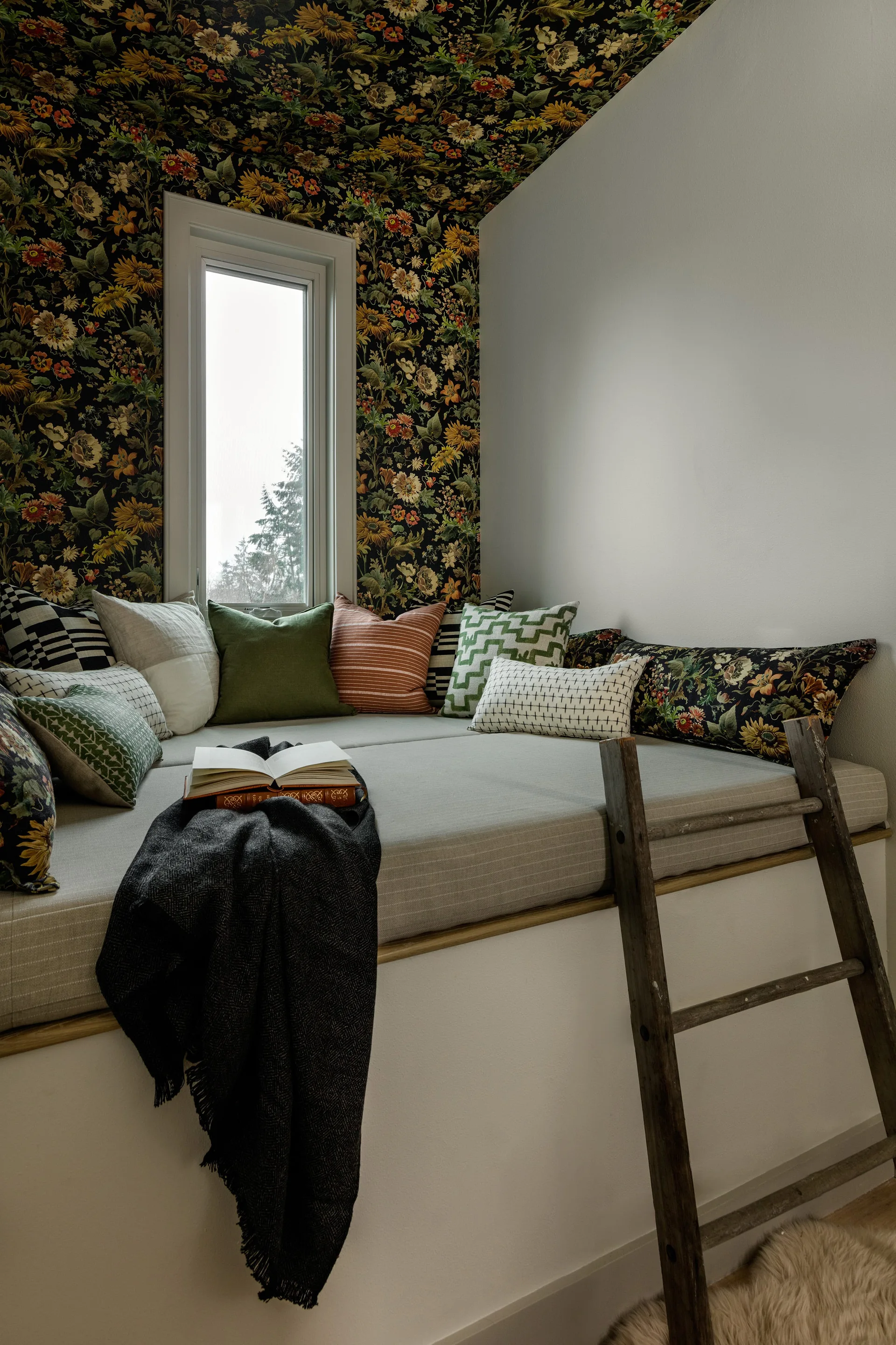 A built-in window seat nook features a floral wallpapered wall and ceiling, several patterned throw pillows, and a rustic wooden ladder.