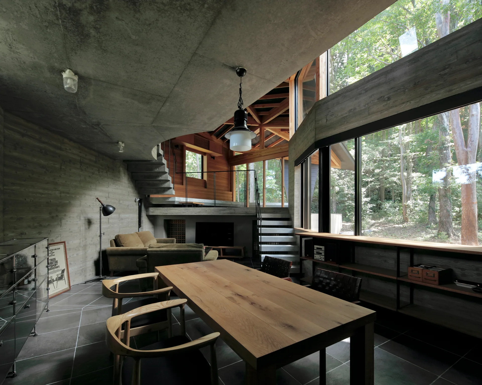 A modern open-concept dining room features a long wooden table, a curved concrete ceiling, and large windows looking out onto a dense forest.