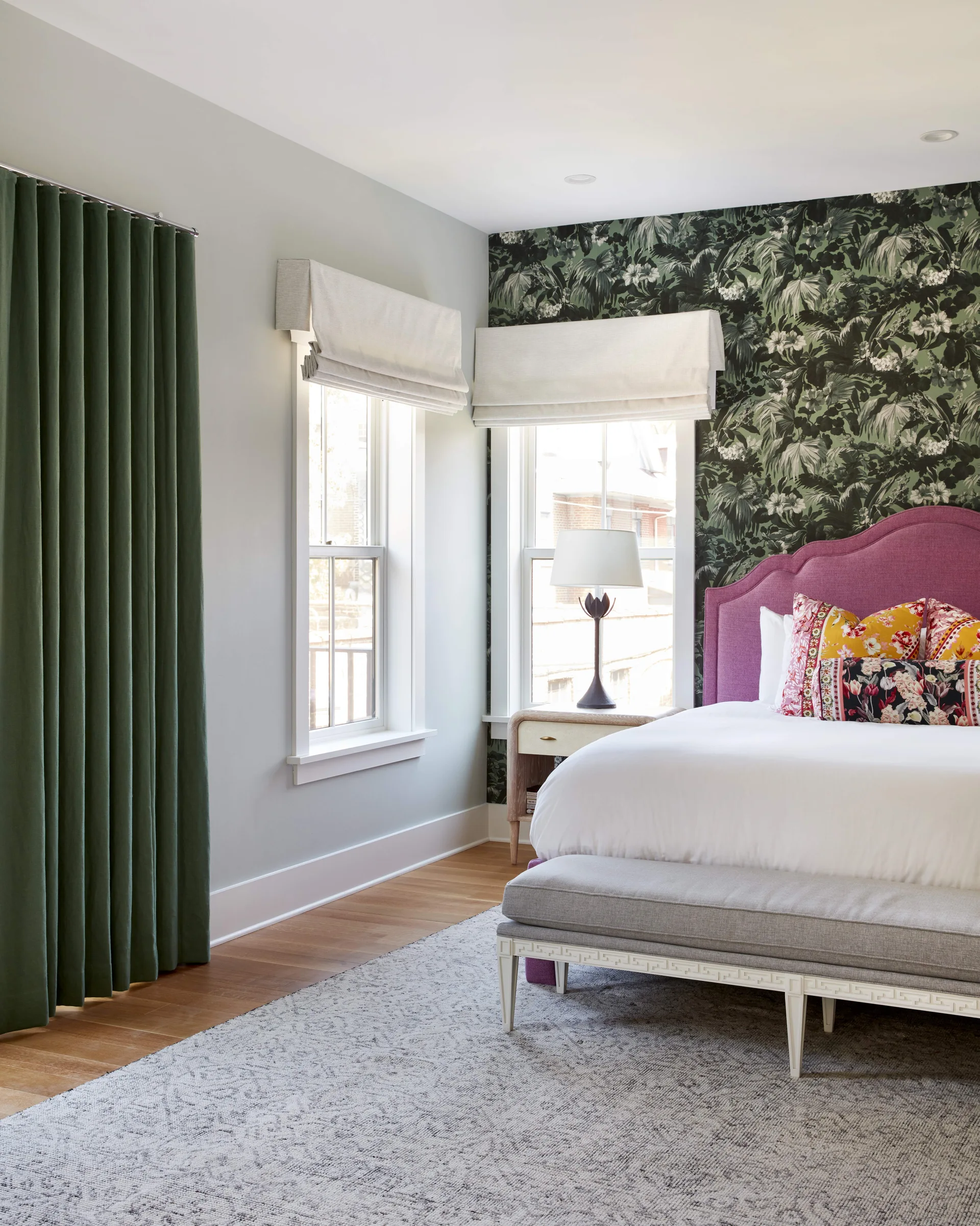 A bedroom shows a purple upholstered headboard, green botanical wallpaper, light grey walls, white roman shades, and a grey bench.