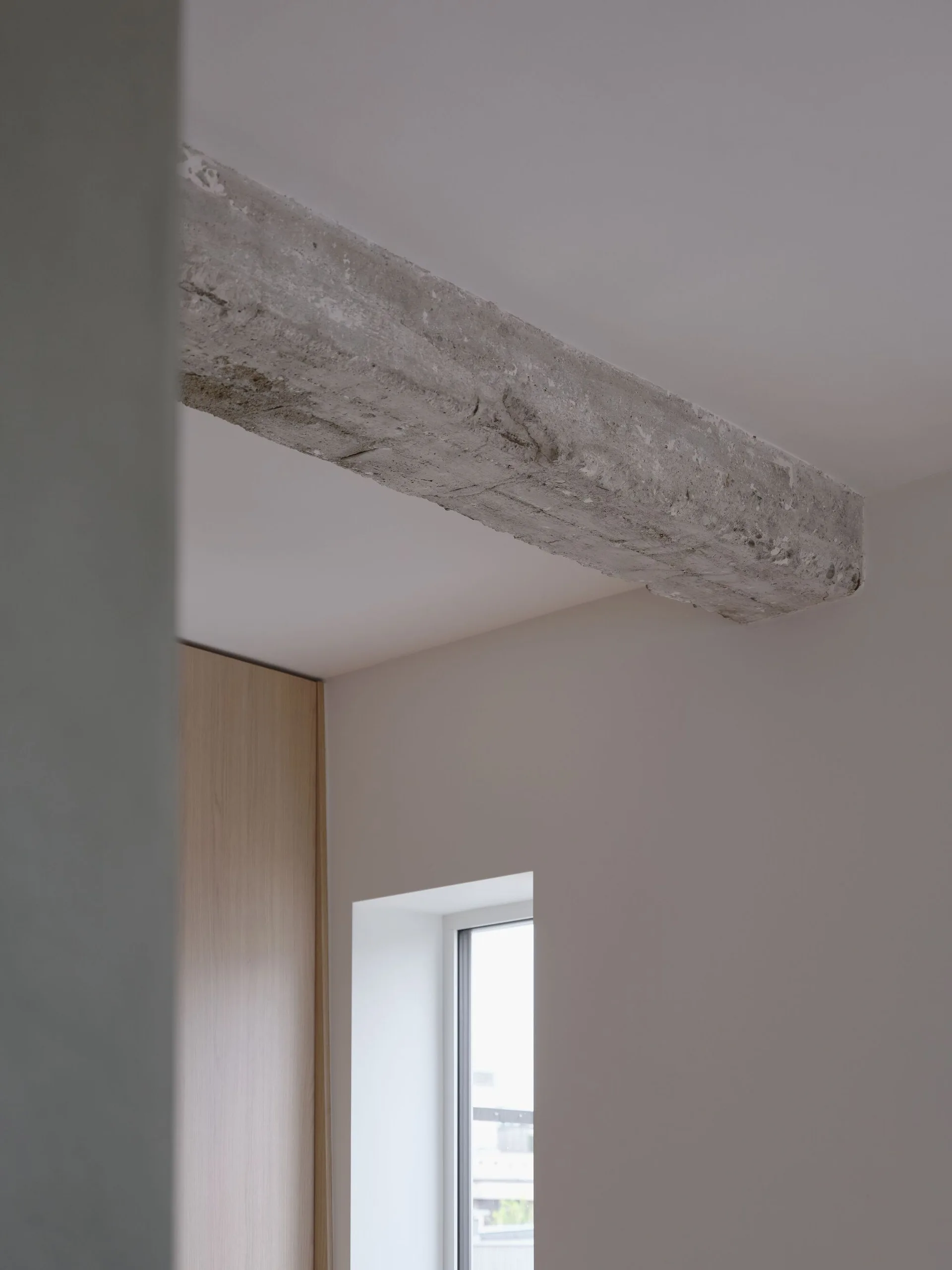 A raw grey concrete ceiling beam juxtaposed with smooth white walls, an inset window, and light wood cabinetry.