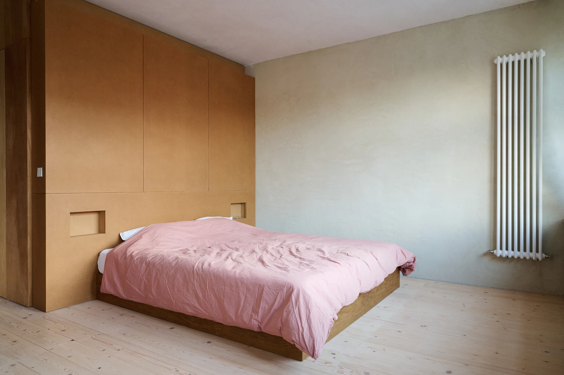 A minimalist bedroom features a low wooden bed with a pink duvet set against a large tan paneled headboard wall with built-in niches.