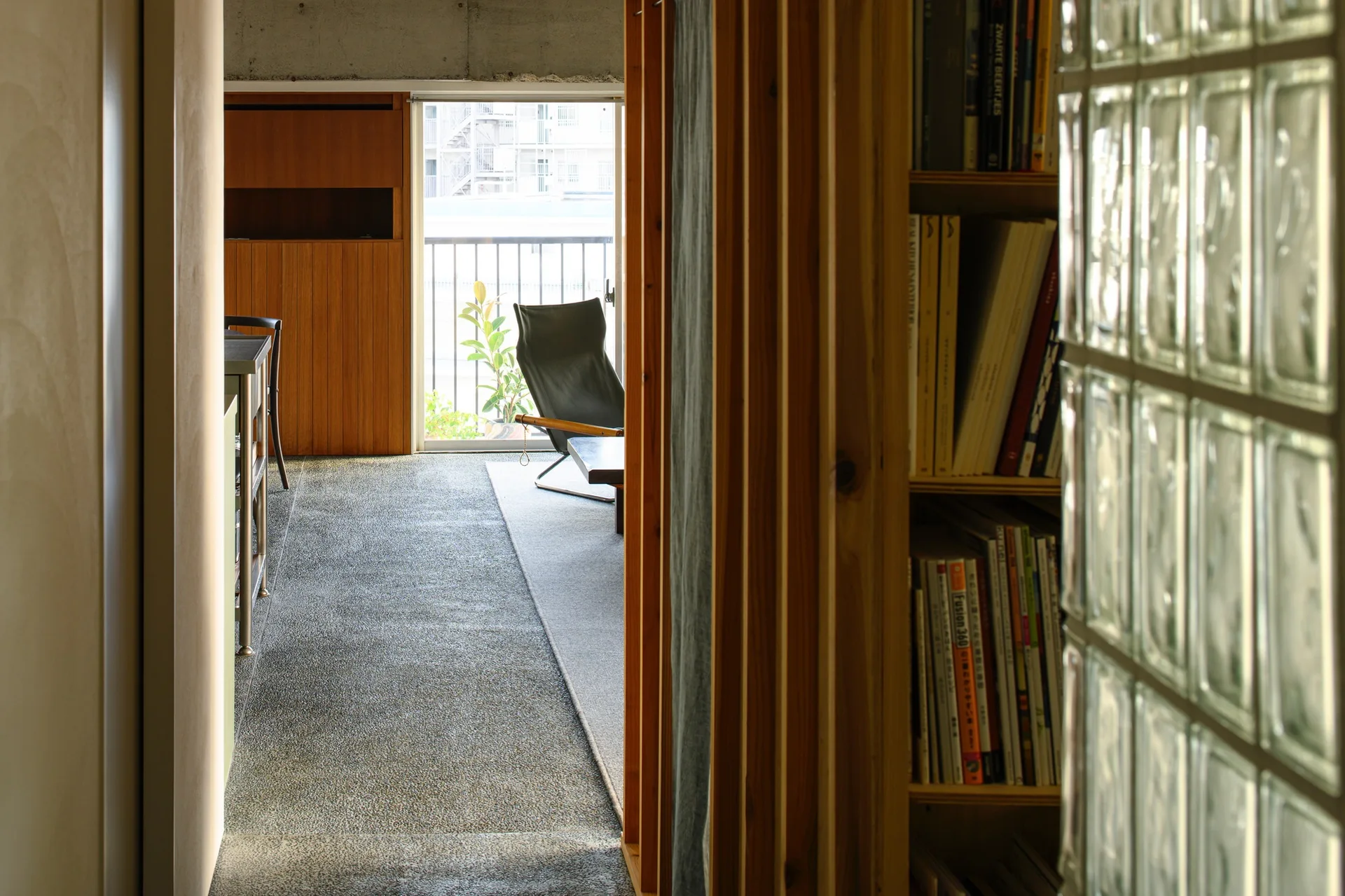 A view down an apartment hallway featuring a wooden bookshelf and glass block wall, leading to a sunlit living room with a chair by the balcony.