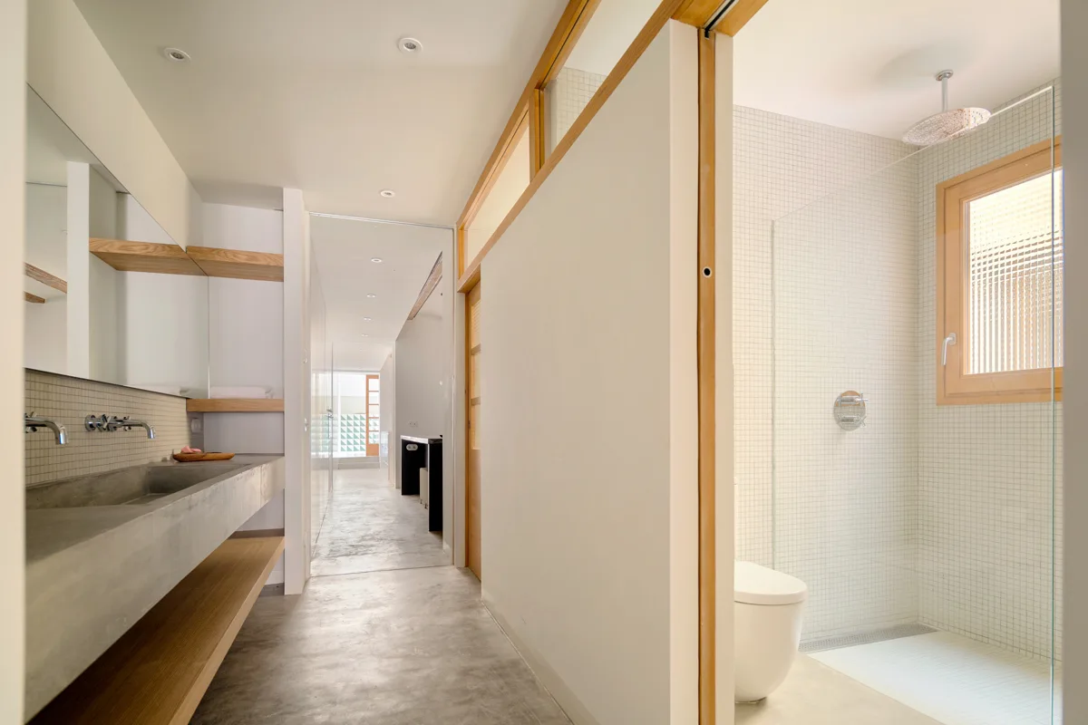 A minimalist long bathroom with a gray concrete trough sink, floating wood shelves, and a walk-in shower with white mosaic tiles.