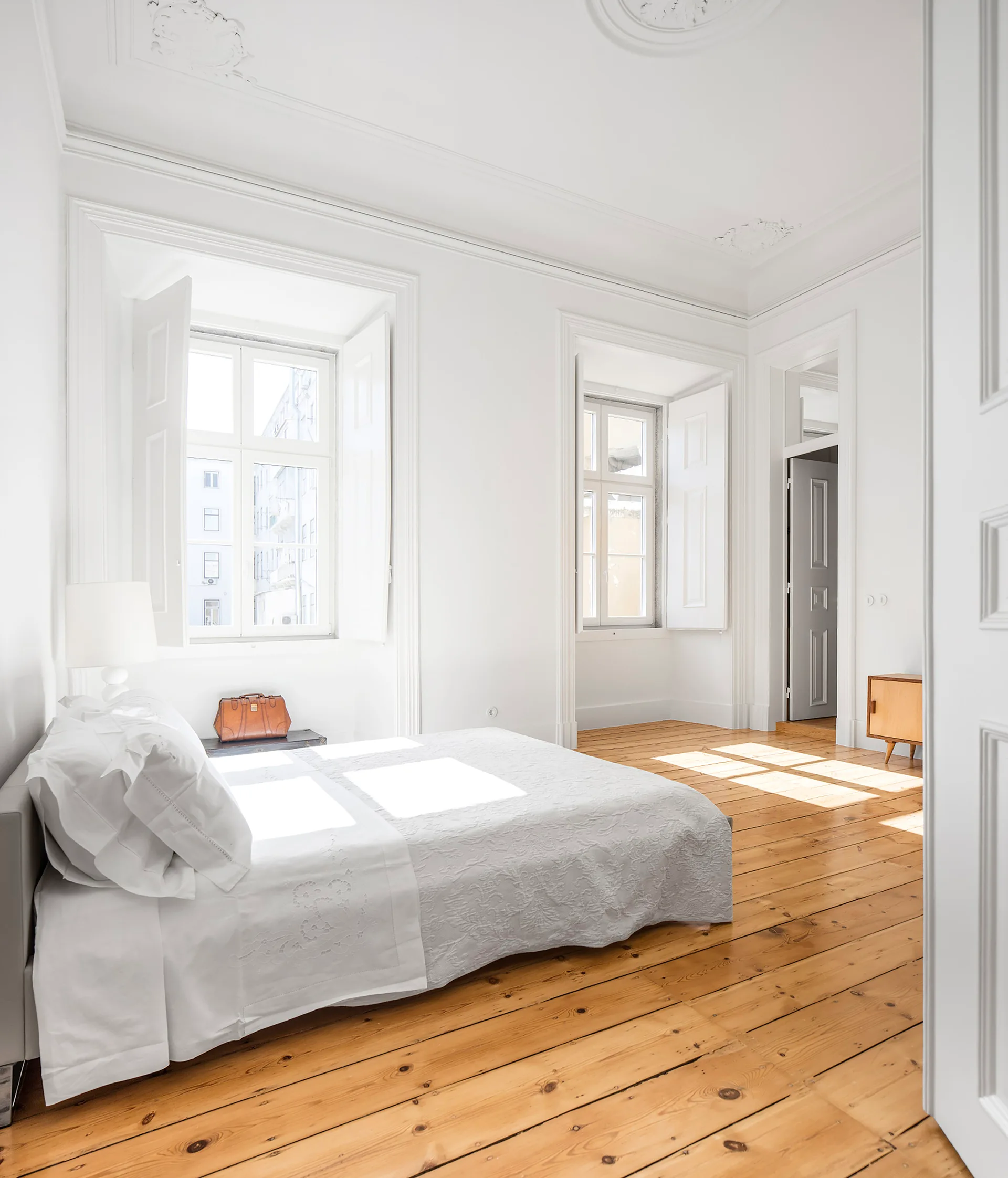 A white bedroom featuring a bed with textured white bedding, wide-plank wood floors, and tall windows with white wood shutters.