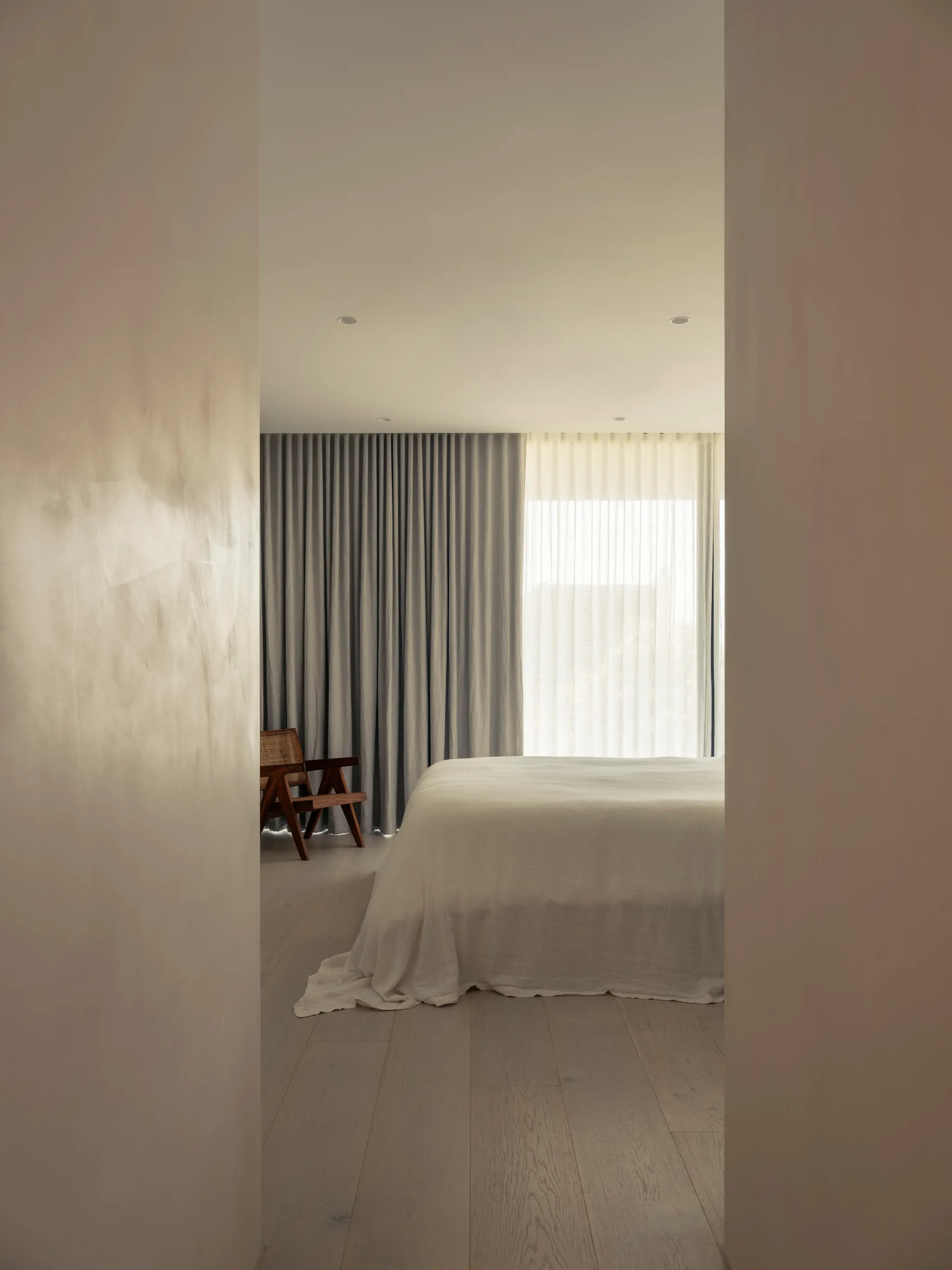 A minimalist bedroom viewed through a doorway, featuring a white linen-covered bed, grey drapes, and a cane accent chair on light wood floors.