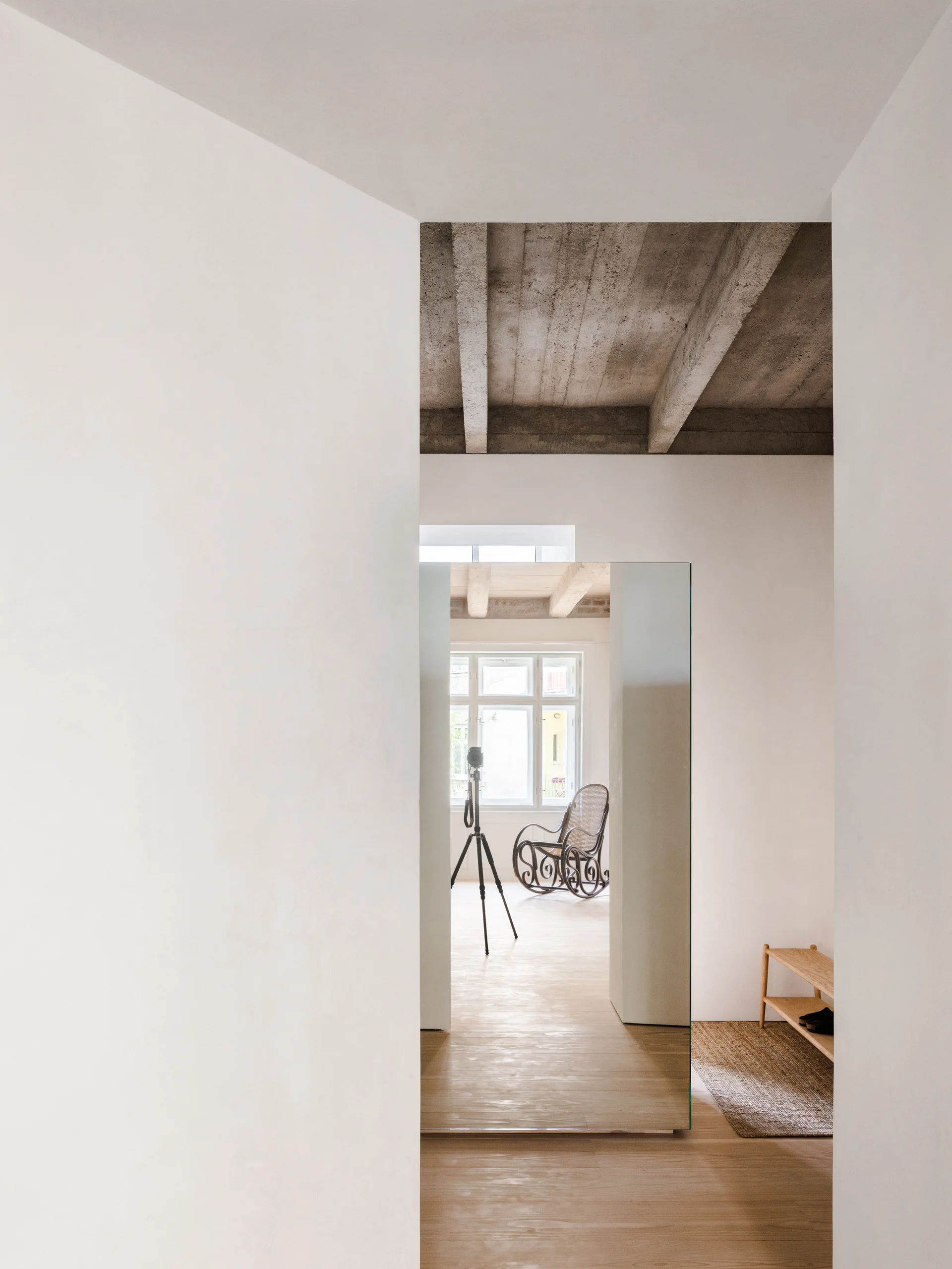 A minimalist hallway with light wood floors and white walls features a large floor mirror reflecting a bright room with a rocking chair.