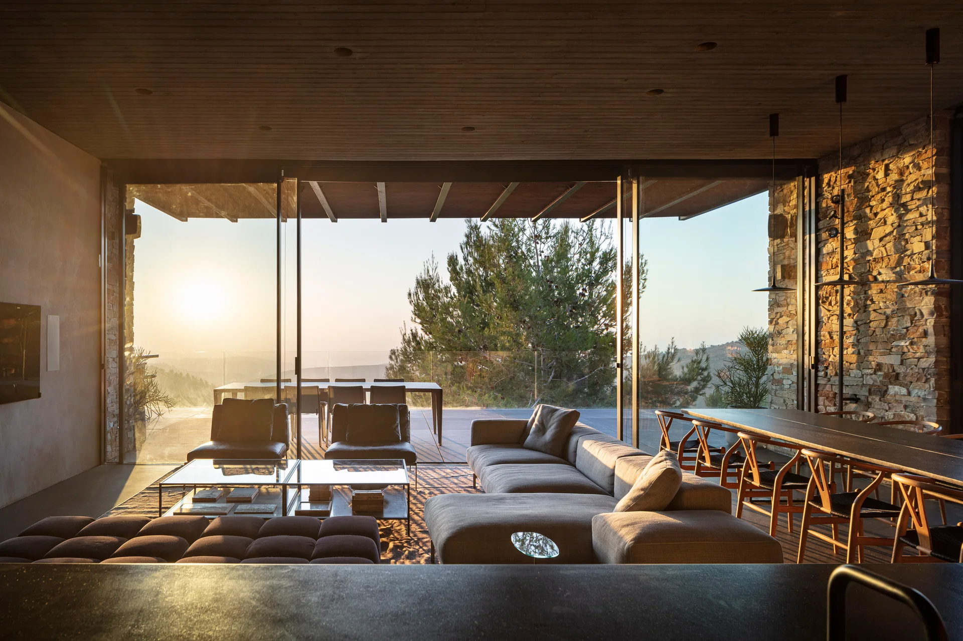 A contemporary living room featuring a grey modular sofa, a stone accent wall, and sliding glass doors that open to an outdoor terrace during sunset.