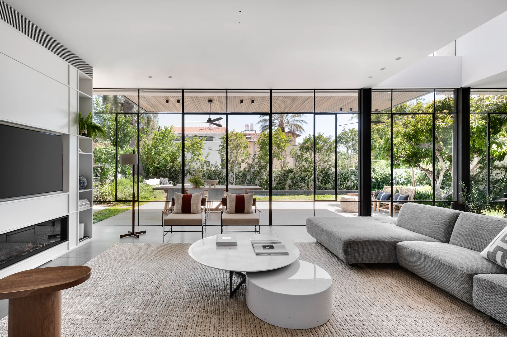 A modern living room features a grey sectional sofa, nesting coffee tables, and a large floor-to-ceiling glass wall overlooking an outdoor patio.
