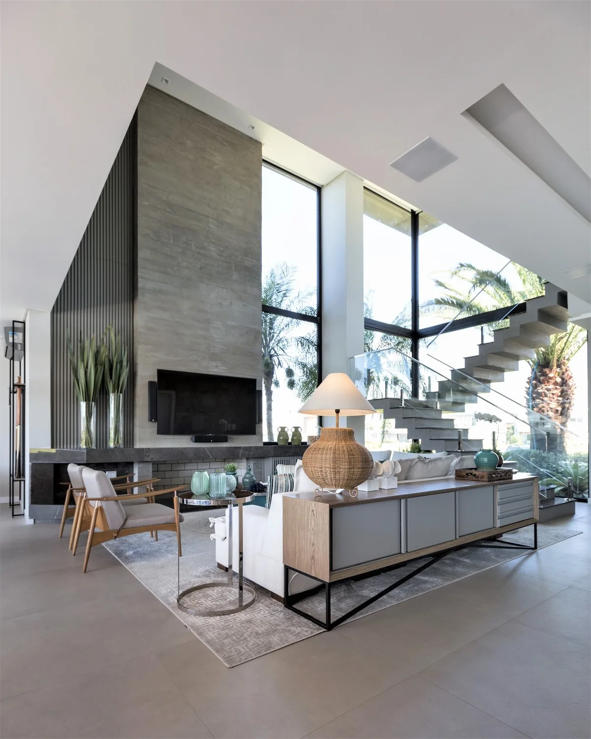 A spacious double-height living room featuring a stone-clad fireplace wall, a white sofa with a console table, and a concrete floating staircase.