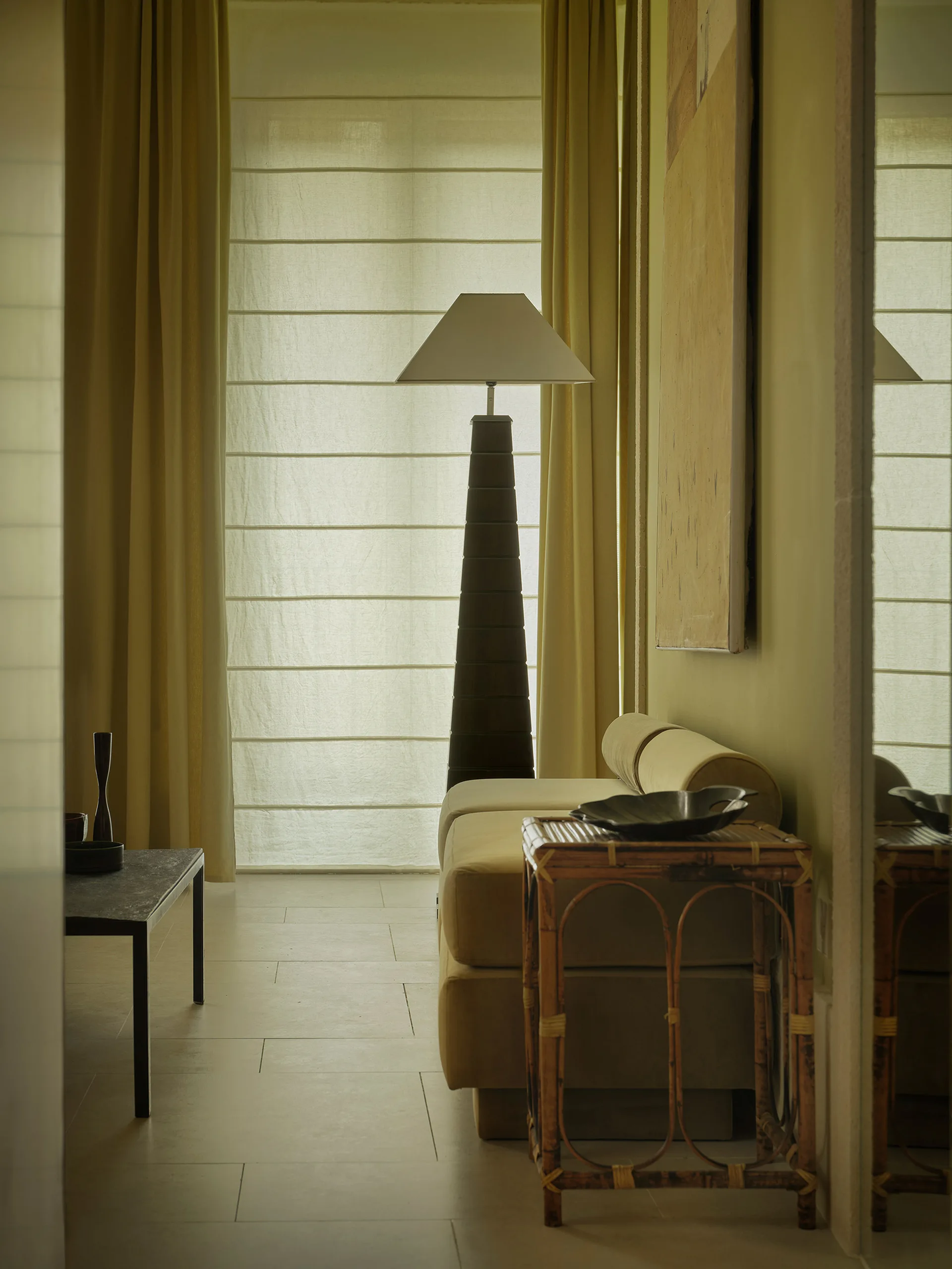 A living room corner features a low beige sofa, a tall tapered floor lamp, a rattan side table, and a fabric roman shade.
