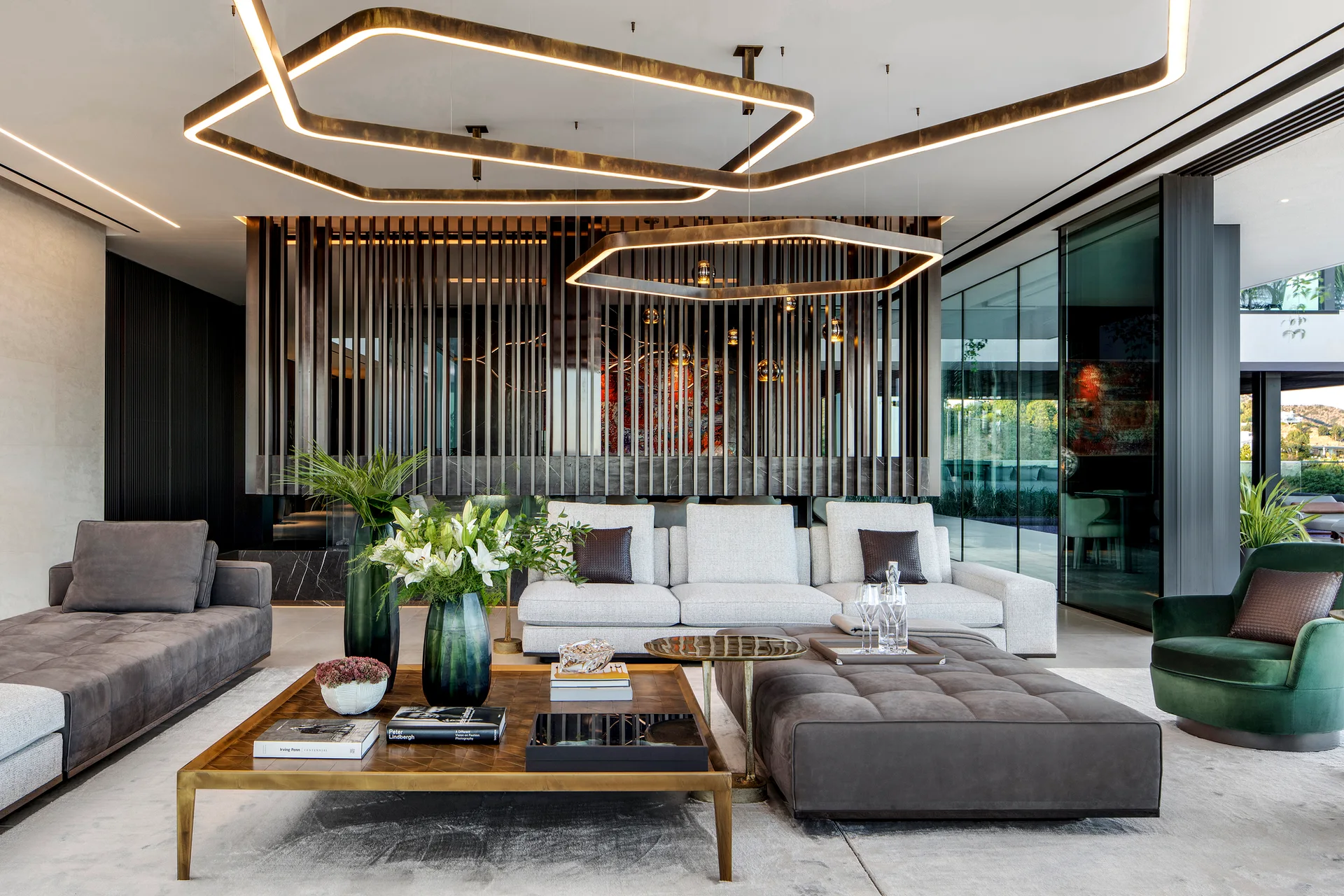 A modern living room features a white sectional sofa, a tufted grey ottoman, a wood slat room divider, and hanging geometric bronze pendant lights.
