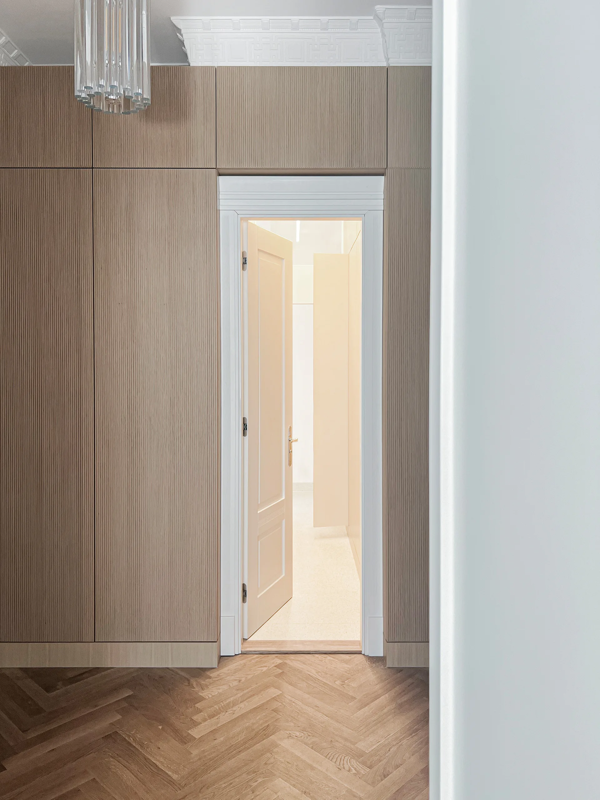 A hallway featuring fluted wood cabinetry surrounding an open white door, set over a light brown herringbone patterned wood floor.
