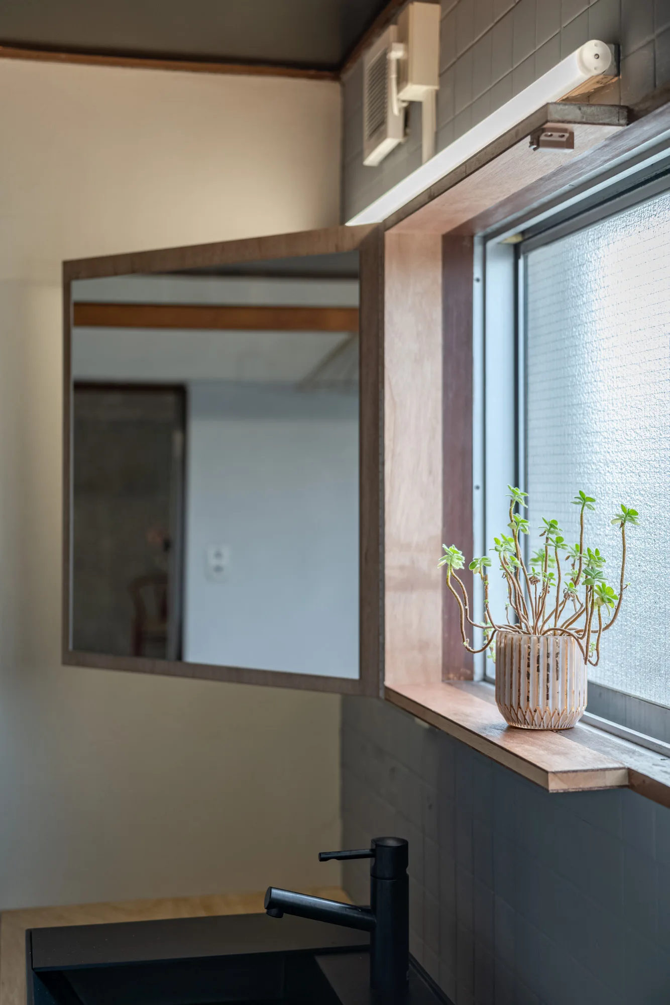 A close-up of a bathroom vanity showing a black faucet, a pivoting wood-framed mirror, and a small potted plant on a wooden window sill.