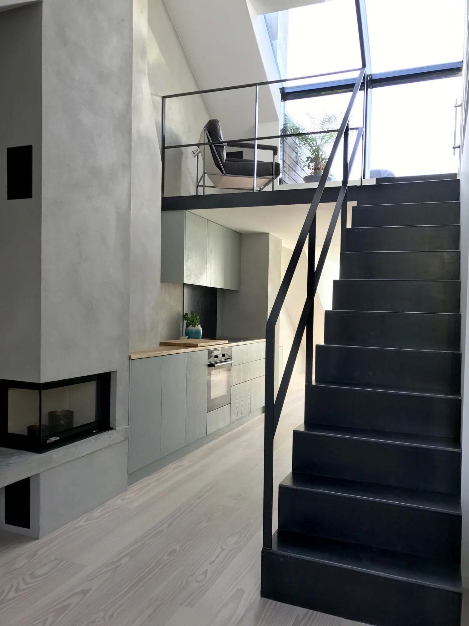 A minimalist loft interior features a black metal staircase, sage green kitchen cabinets under a mezzanine, and a skylight.