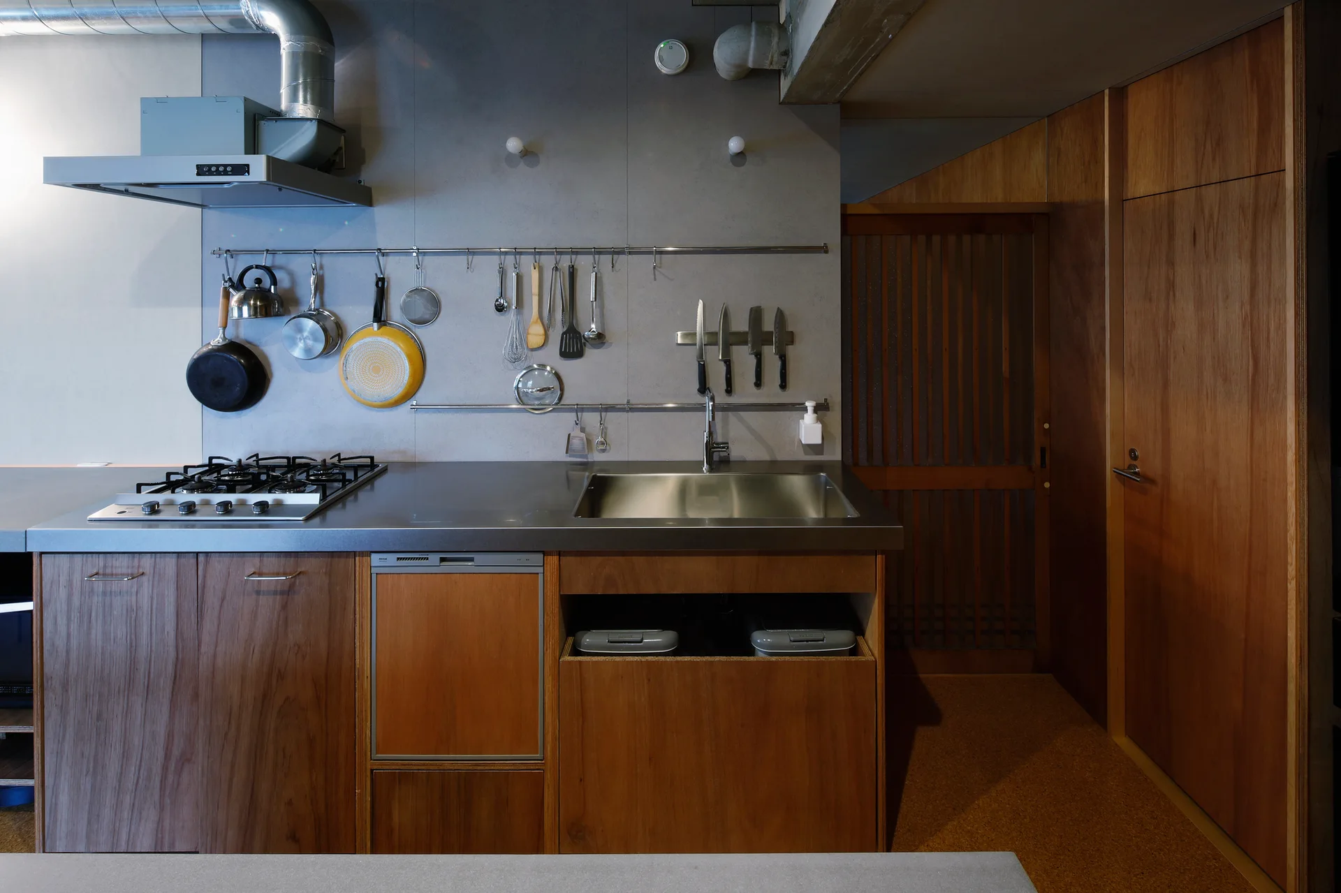 A modern industrial kitchen features a stainless steel countertop, wood-toned lower cabinets, a gas stove, and pots hanging from a wall-mounted rail.