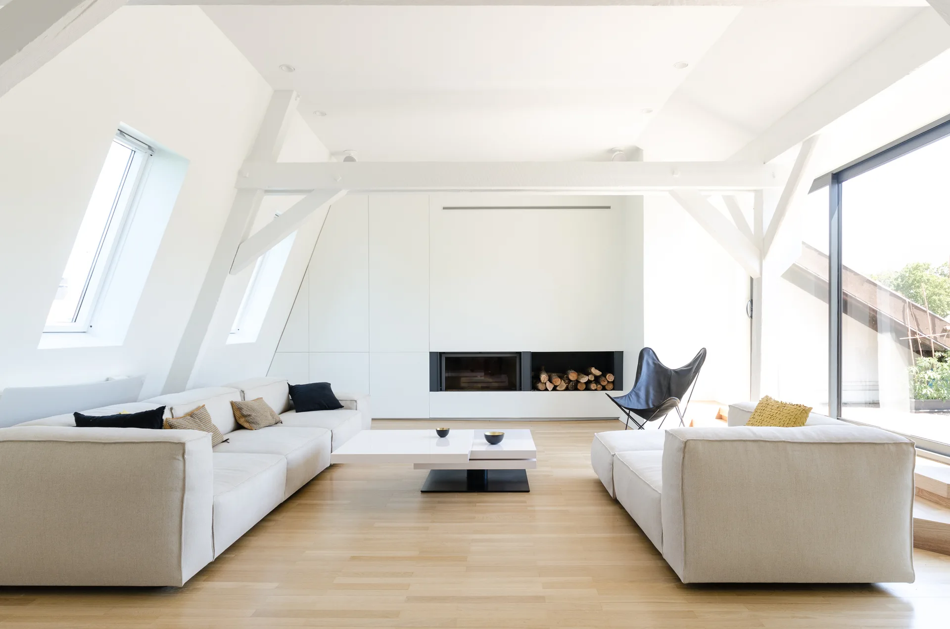 A bright attic living room features two off-white sofas, a low coffee table, exposed white ceiling beams, and a built-in fireplace.