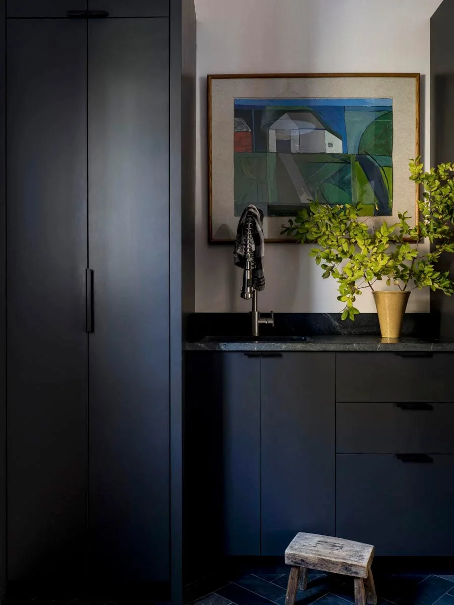 Charcoal grey built-in cabinetry frames a utility sink, topped by framed artwork and a vase of green branches, with a small wooden stool on the floor.
