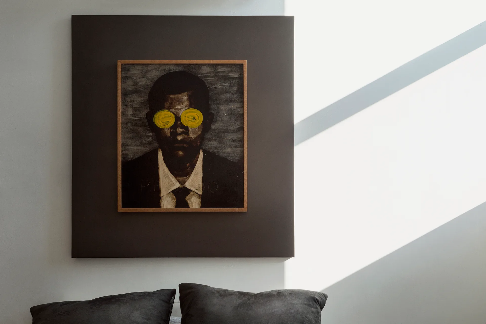 A framed portrait painting with yellow paint over the eyes hangs on a dark grey square panel above dark pillows in a minimalist room.