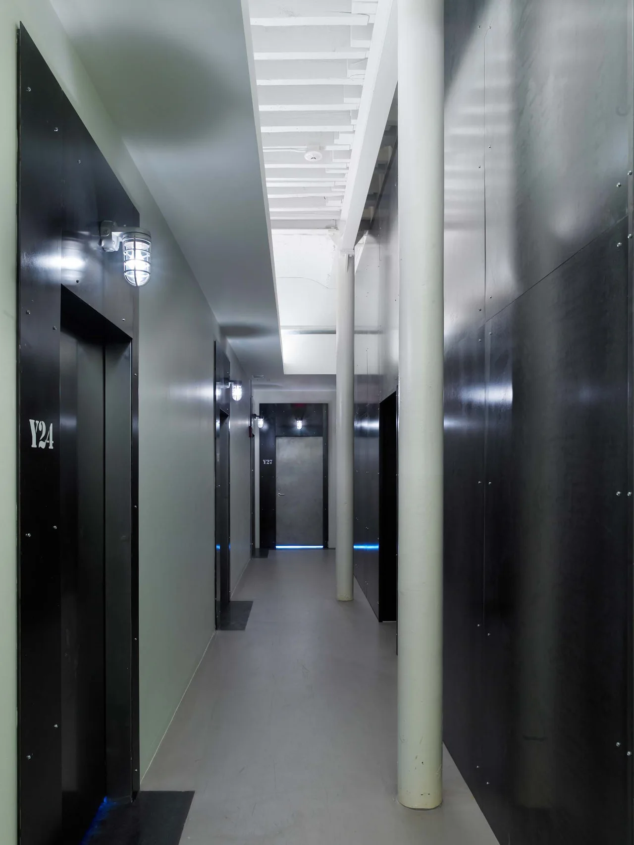 A long industrial hallway features dark metal wall panels, structural white columns, an exposed ceiling, and caged wall sconces.