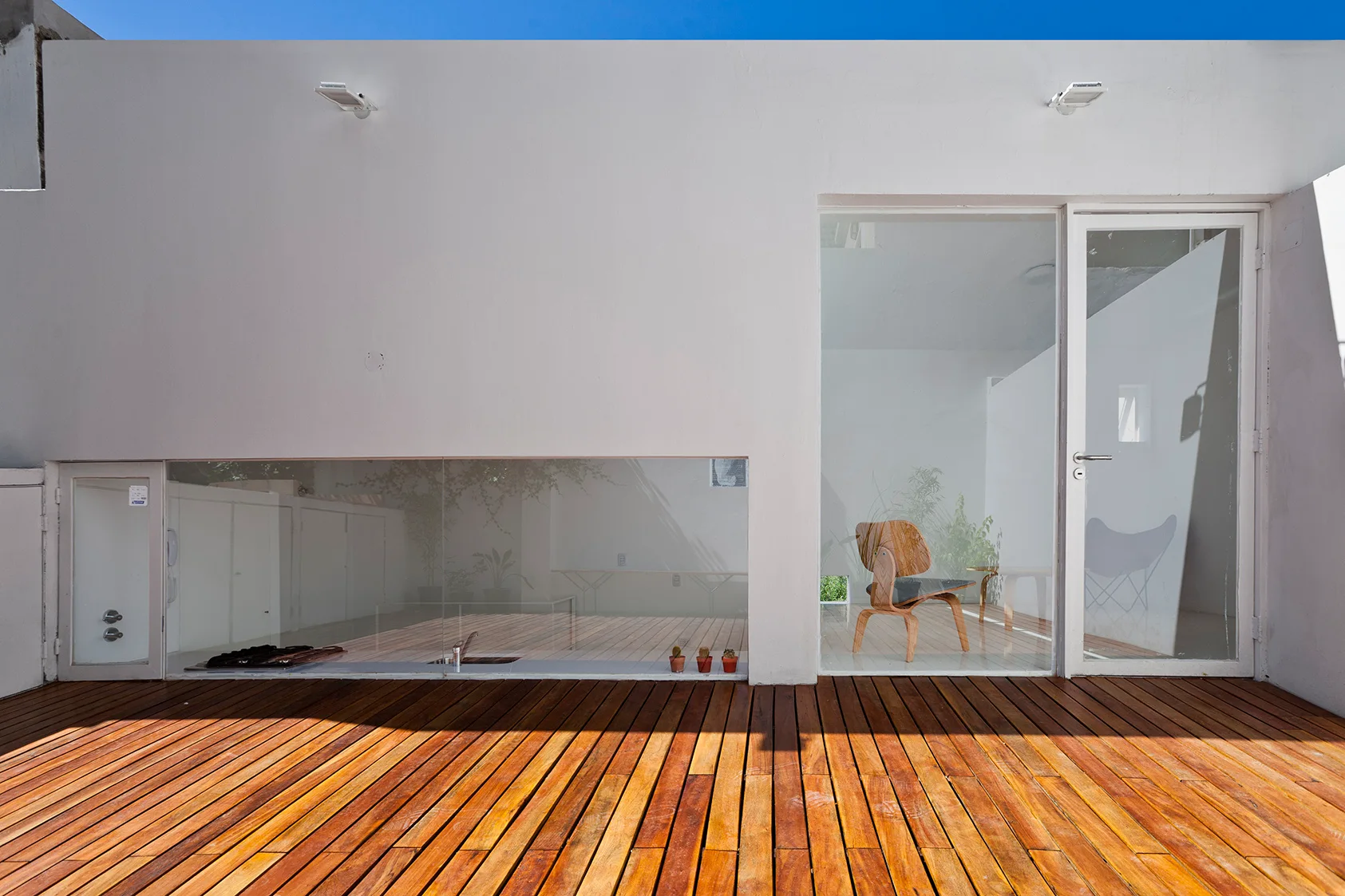 A minimalist outdoor wooden deck leads to a plain white facade with a low-set rectangular window and a glass door revealing a modern chair inside.