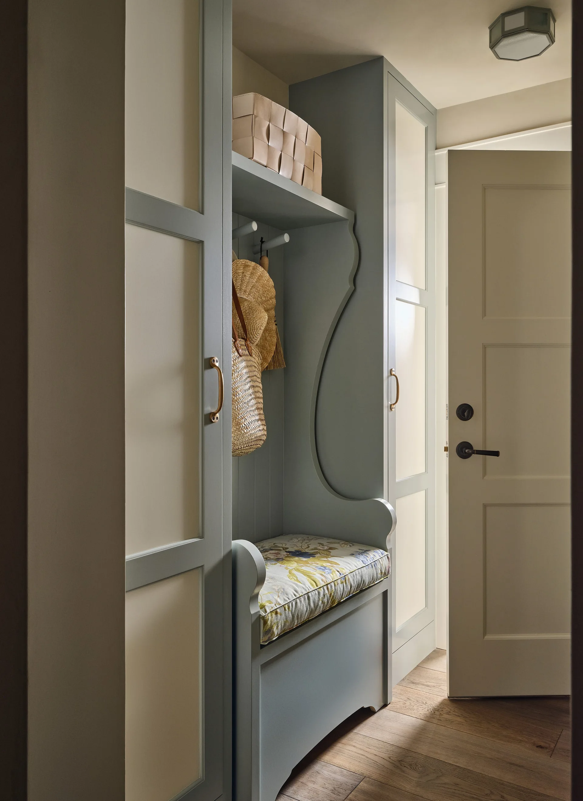 A built-in muted blue mudroom storage bench features a floral print cushion, storage cabinets, and woven baskets next to an open door.