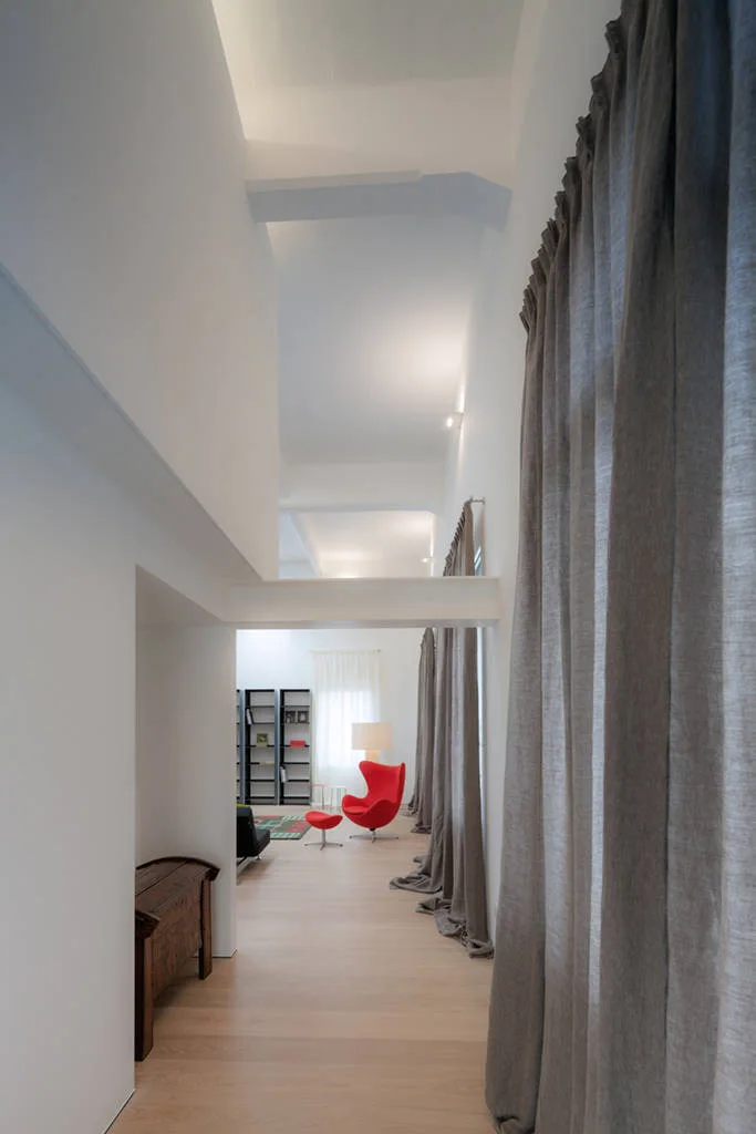 A tall minimalist hallway with light wood floors and grey curtains leads to a seating area with a bright red egg-shaped armchair.