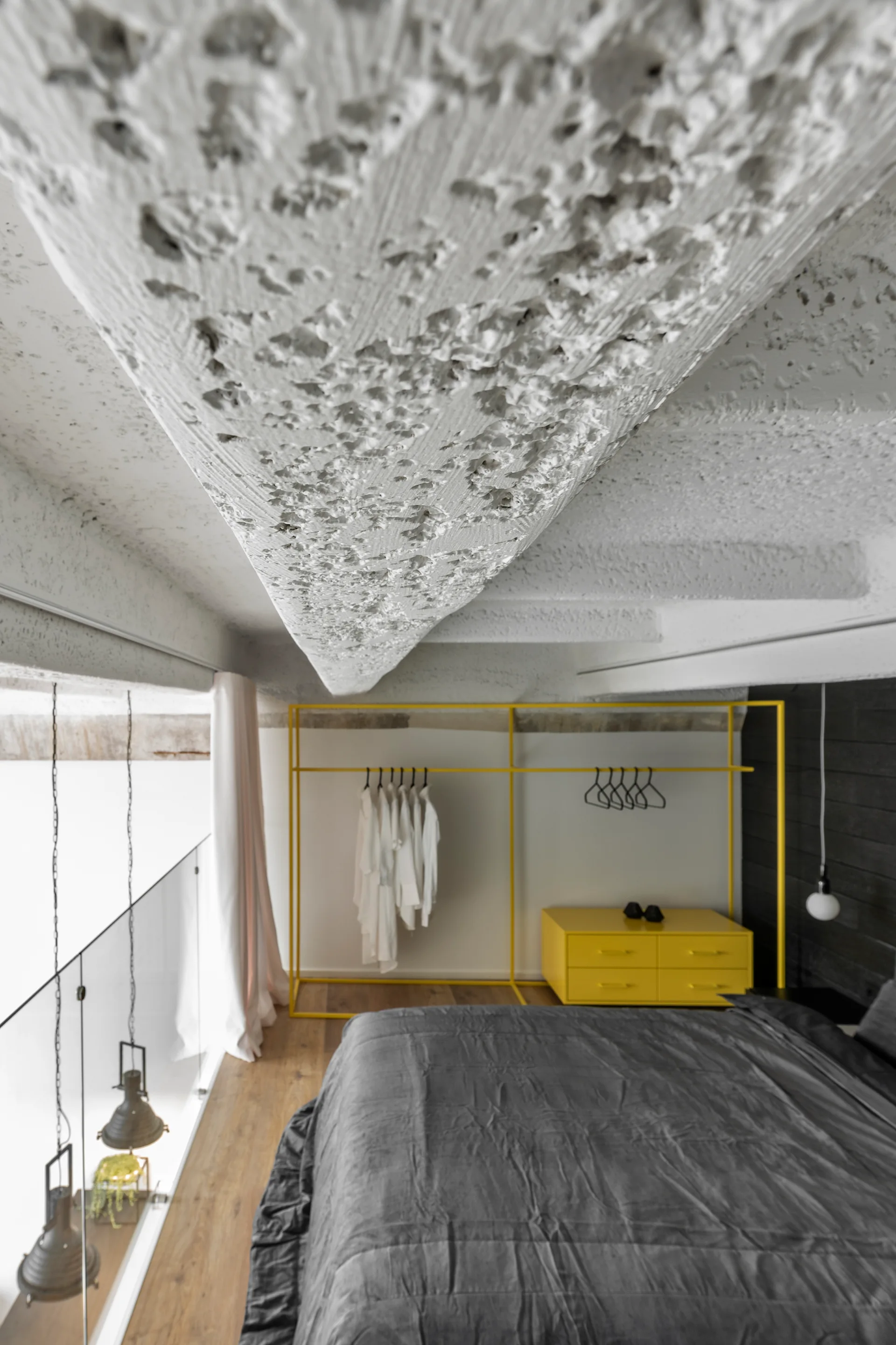A loft bedroom features a rough textured concrete ceiling, a grey bed, and a bright yellow open clothes rack next to a matching dresser.