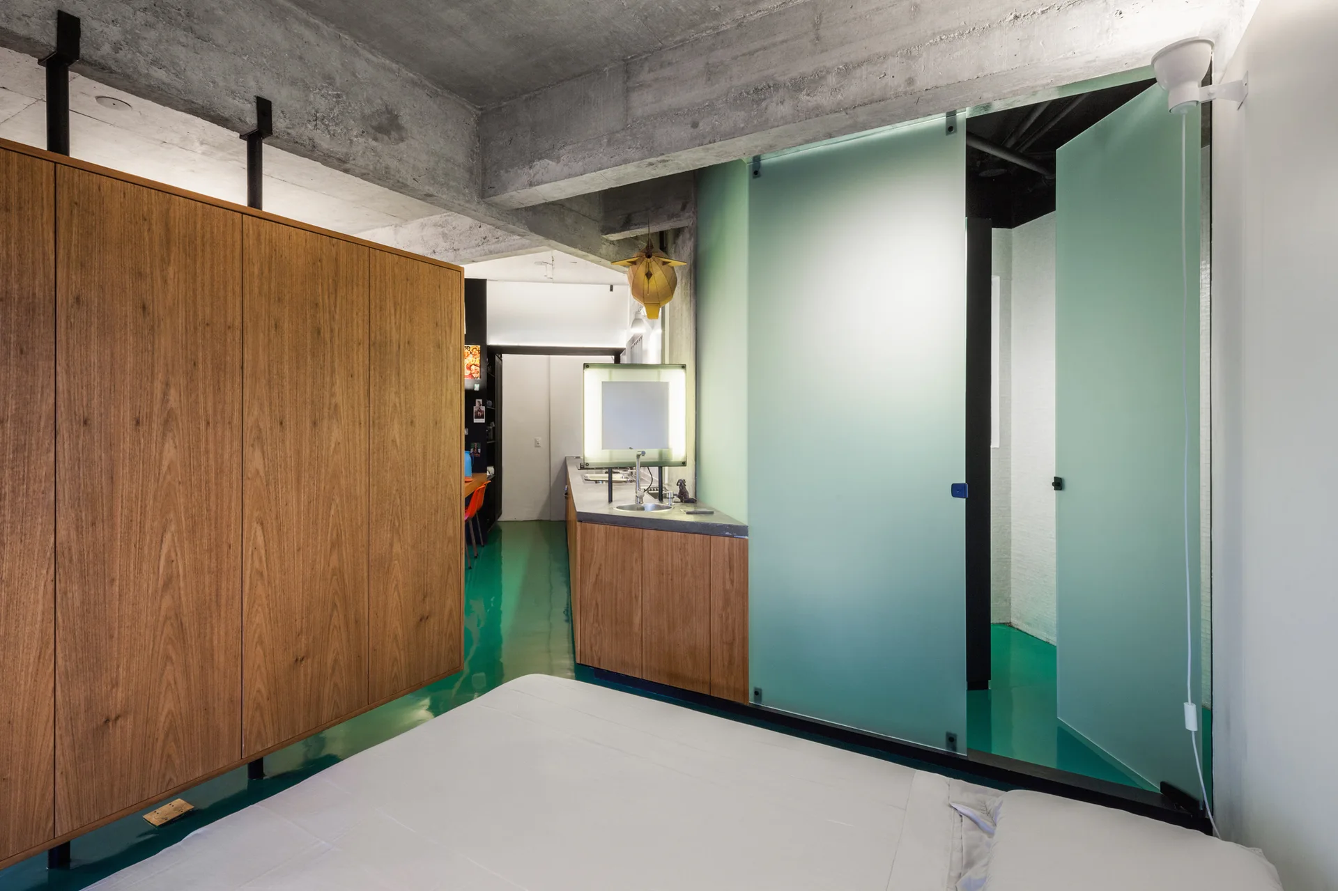 A modern bedroom with wood wardrobes and an exposed concrete ceiling features a glossy green floor and matching frosted glass bathroom doors.