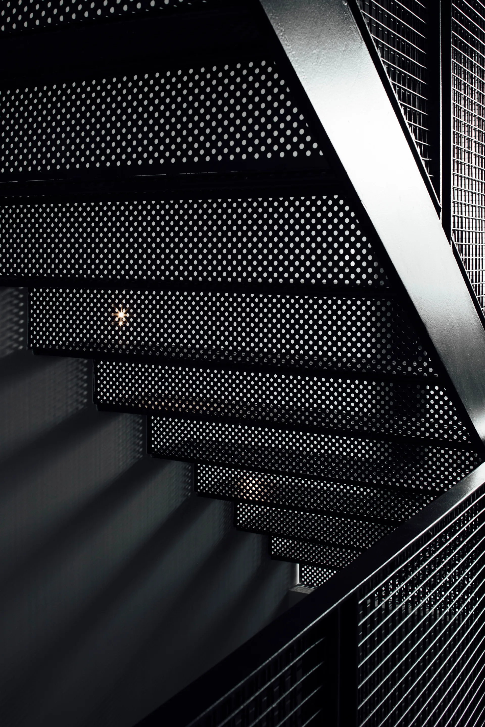 A close-up view of an industrial matte black steel staircase featuring perforated treads and a wire mesh railing against a dark wall.