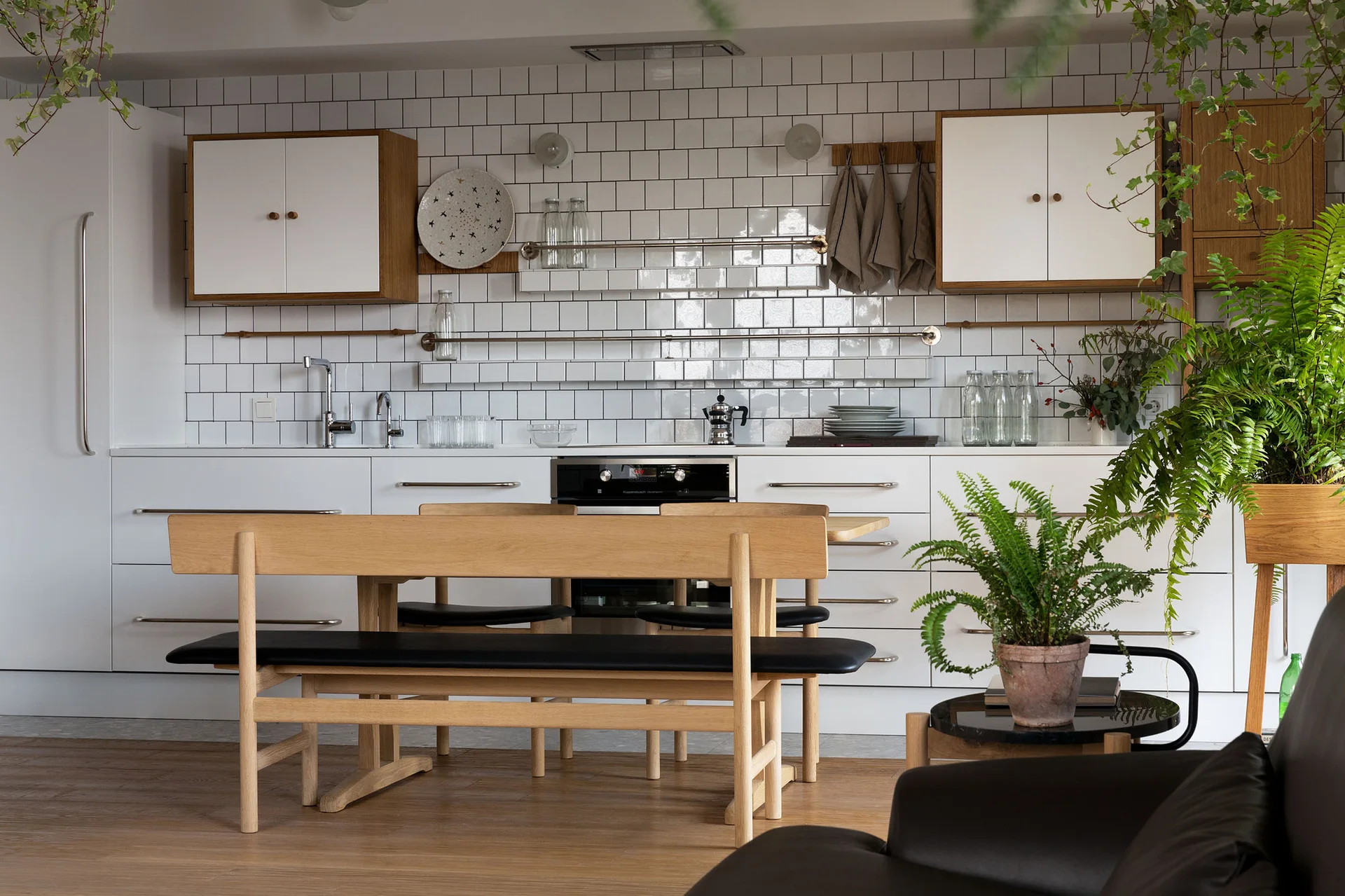 A modern Scandinavian kitchen features white square tile backsplash, wood-trimmed upper cabinets, and a light wood dining bench with a black cushion.