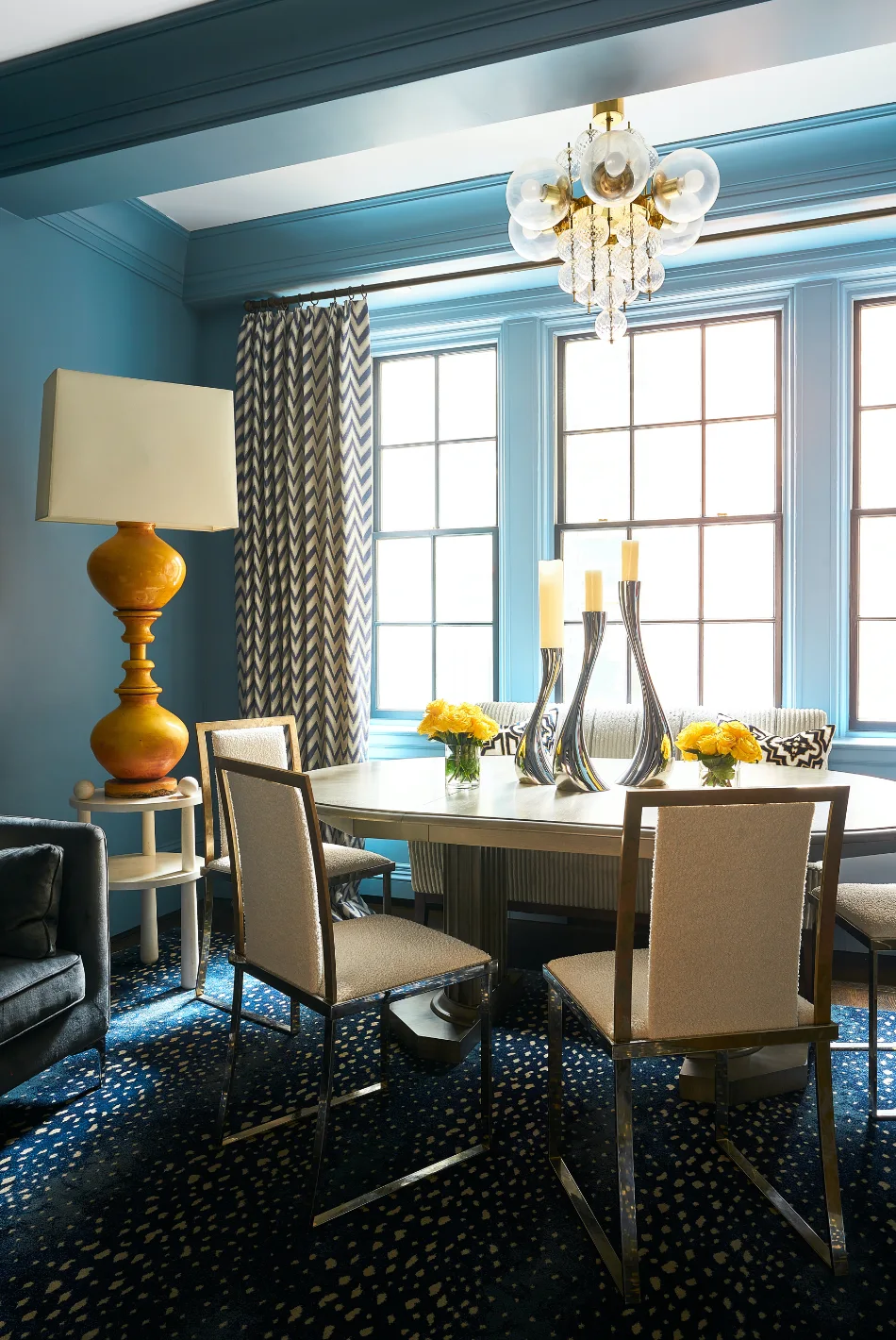 A dining room features blue painted walls and ceiling, a round dining table, upholstered chairs, chevron curtains, and a tall yellow table lamp.
