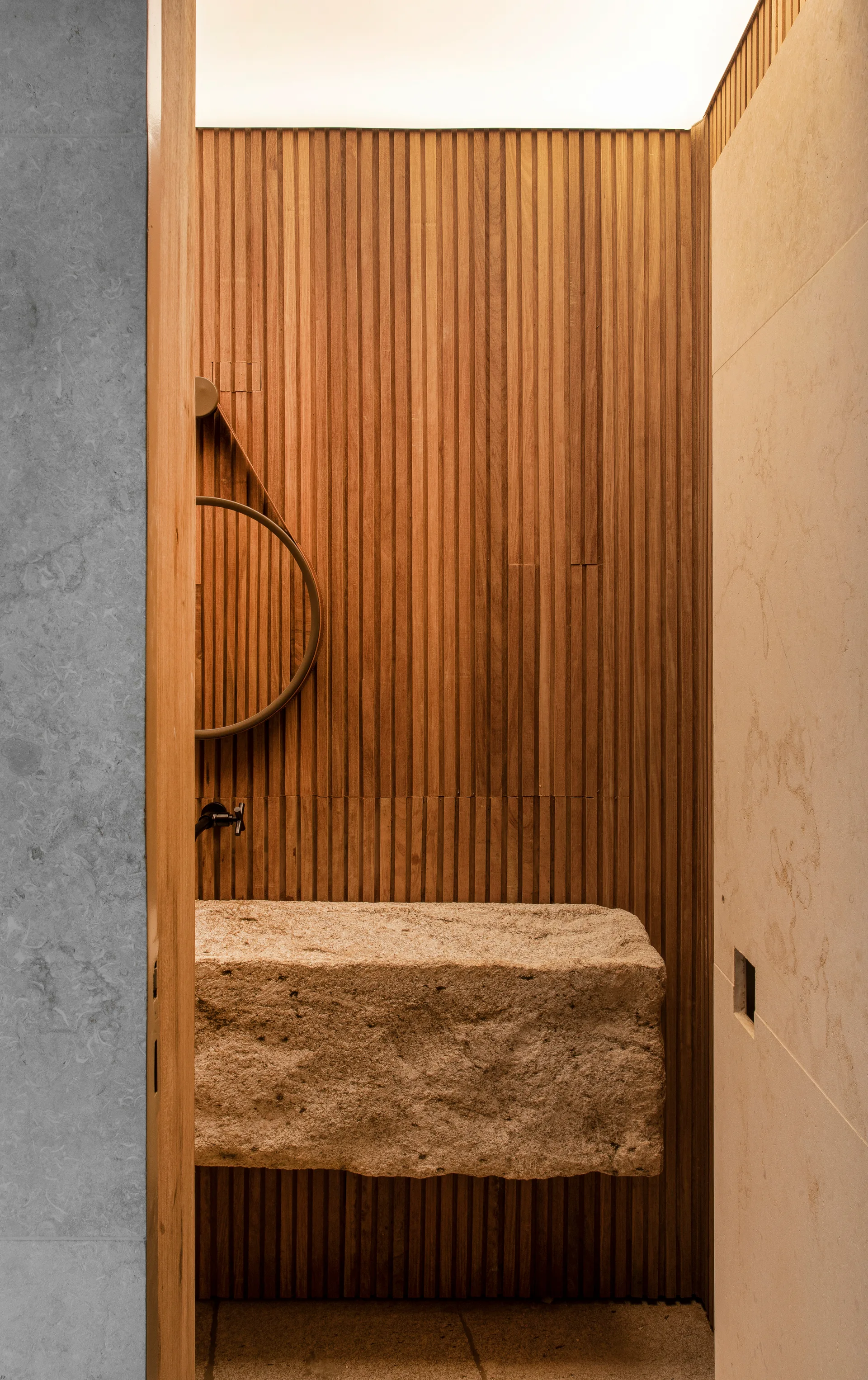 A powder room features a floating rough-hewn stone sink set against a vertical slatted wood wall with a circular hanging mirror.