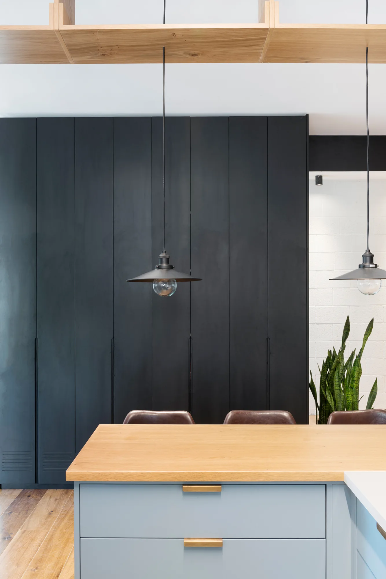 A modern kitchen features a blue-grey island with a wood countertop, black pendant lights, and a full-height wall of matte black cabinetry.