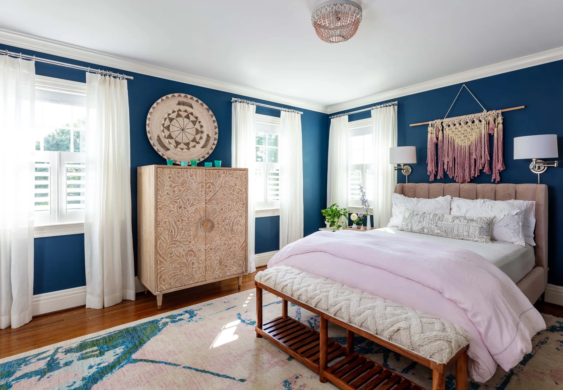 A bedroom features navy blue walls, an upholstered bed with pink bedding, a carved wooden cabinet, and white shuttered windows with sheer curtains.