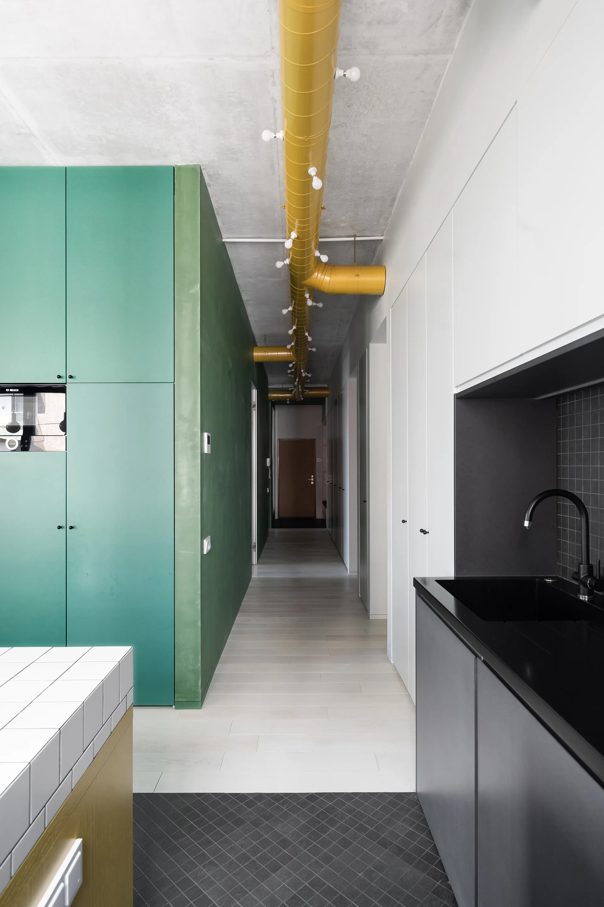 A modern hallway next to a kitchen featuring green cabinets, a concrete ceiling, and a yellow industrial duct pipe with exposed light bulbs.