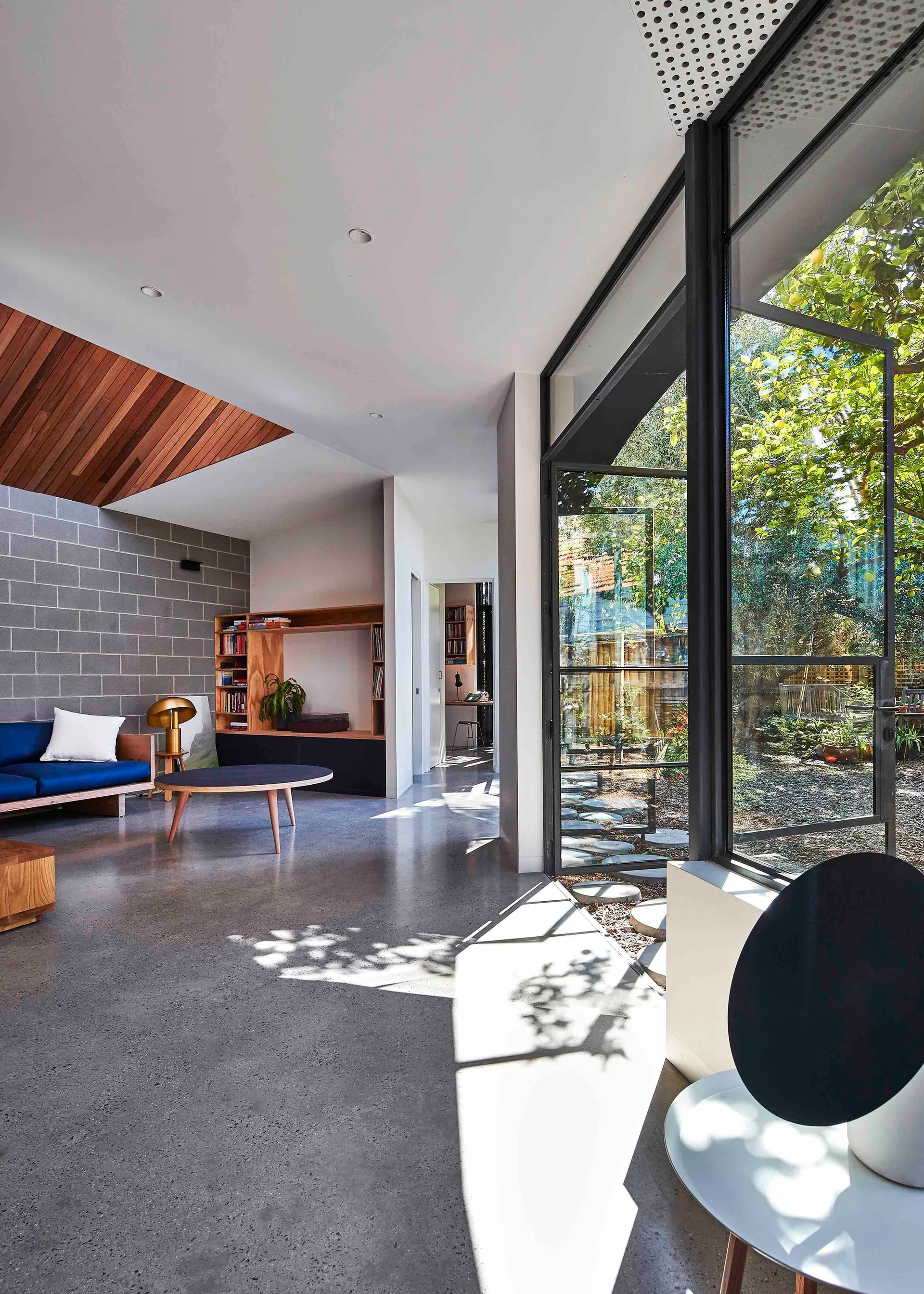 A modern living room with polished concrete flooring, a gray cinder block accent wall, a blue sofa, and large black-framed windows looking out to a garden.