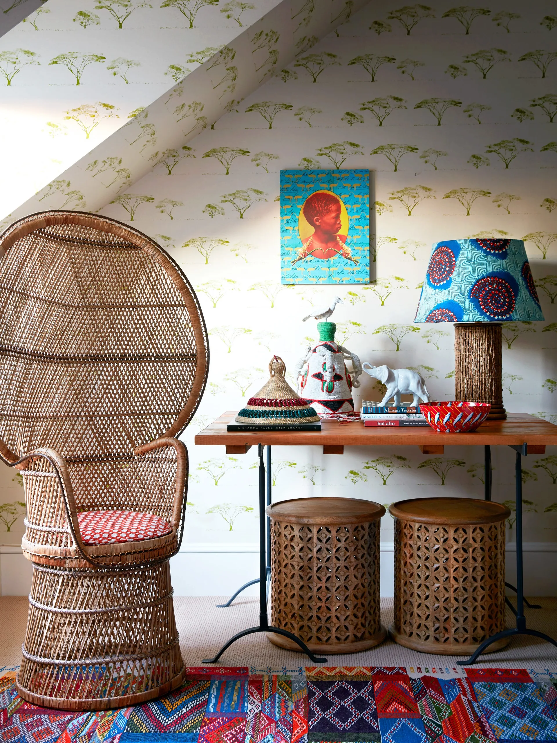 A wicker peacock chair and wooden console table stand against tree-patterned wallpaper, styled with carved wooden stools and a colorful geometric rug.