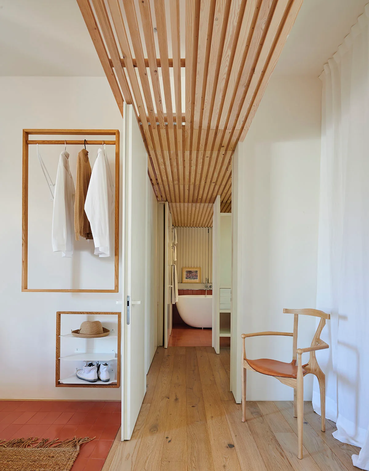 A minimalist dressing corridor features a wood slat ceiling, wall-mounted oak clothing racks, a wooden chair, and a view into a bathroom.