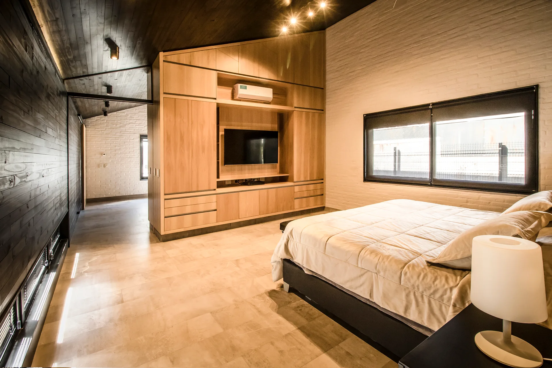 A modern bedroom features a bed with beige bedding, a large light-wood built-in wardrobe with an integrated TV, and a slanted dark wood ceiling.