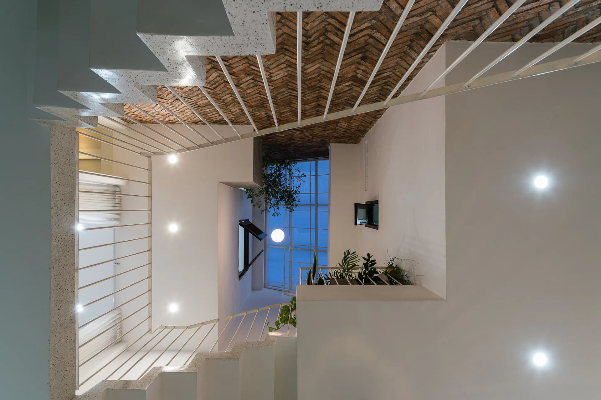 A vertical view looking up a white minimalist staircase with thin metal railings toward an exposed brick herringbone ceiling.