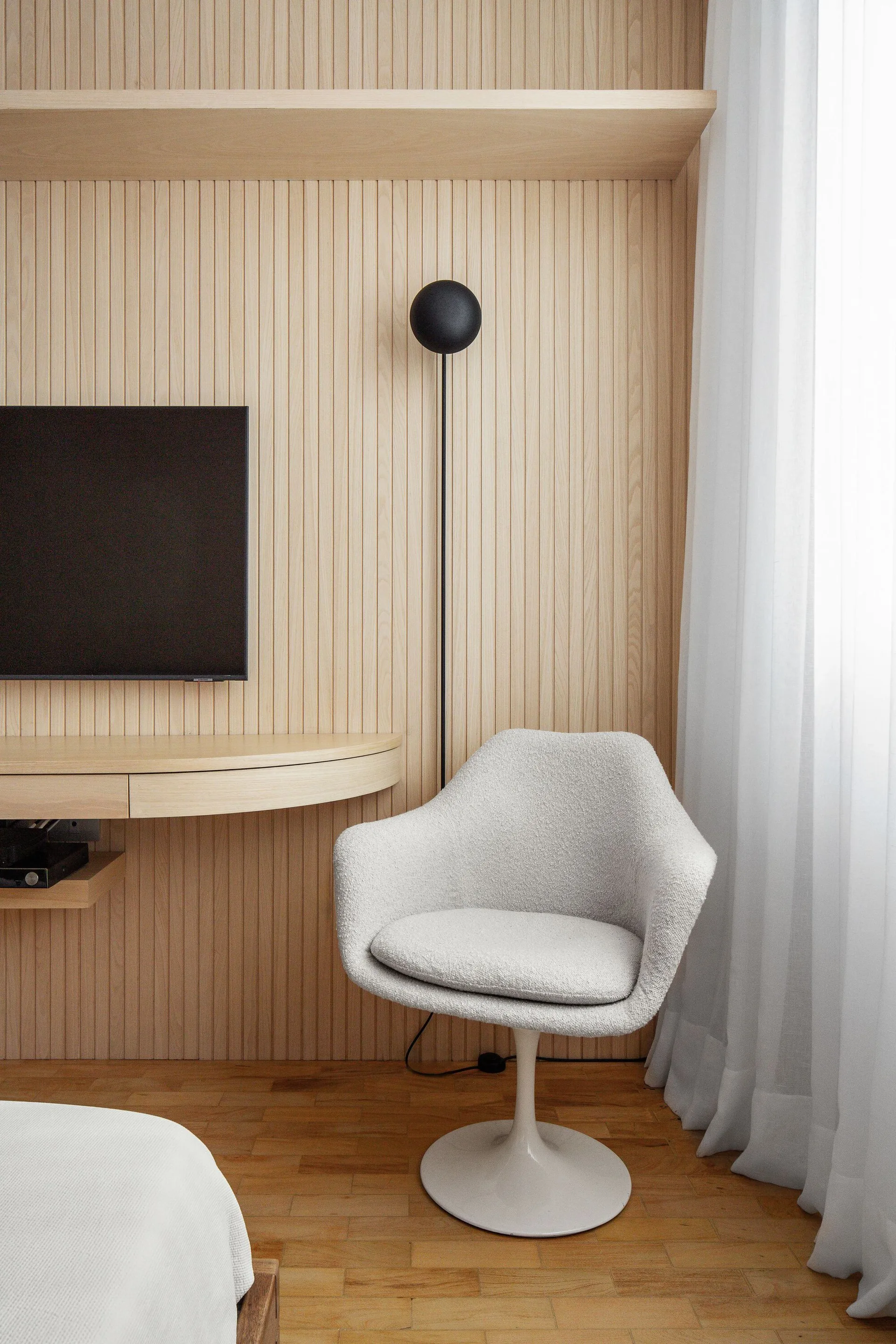 A white tulip chair sits next to a curved wood floating desk against a vertical wood slat accent wall in a minimalist bedroom.