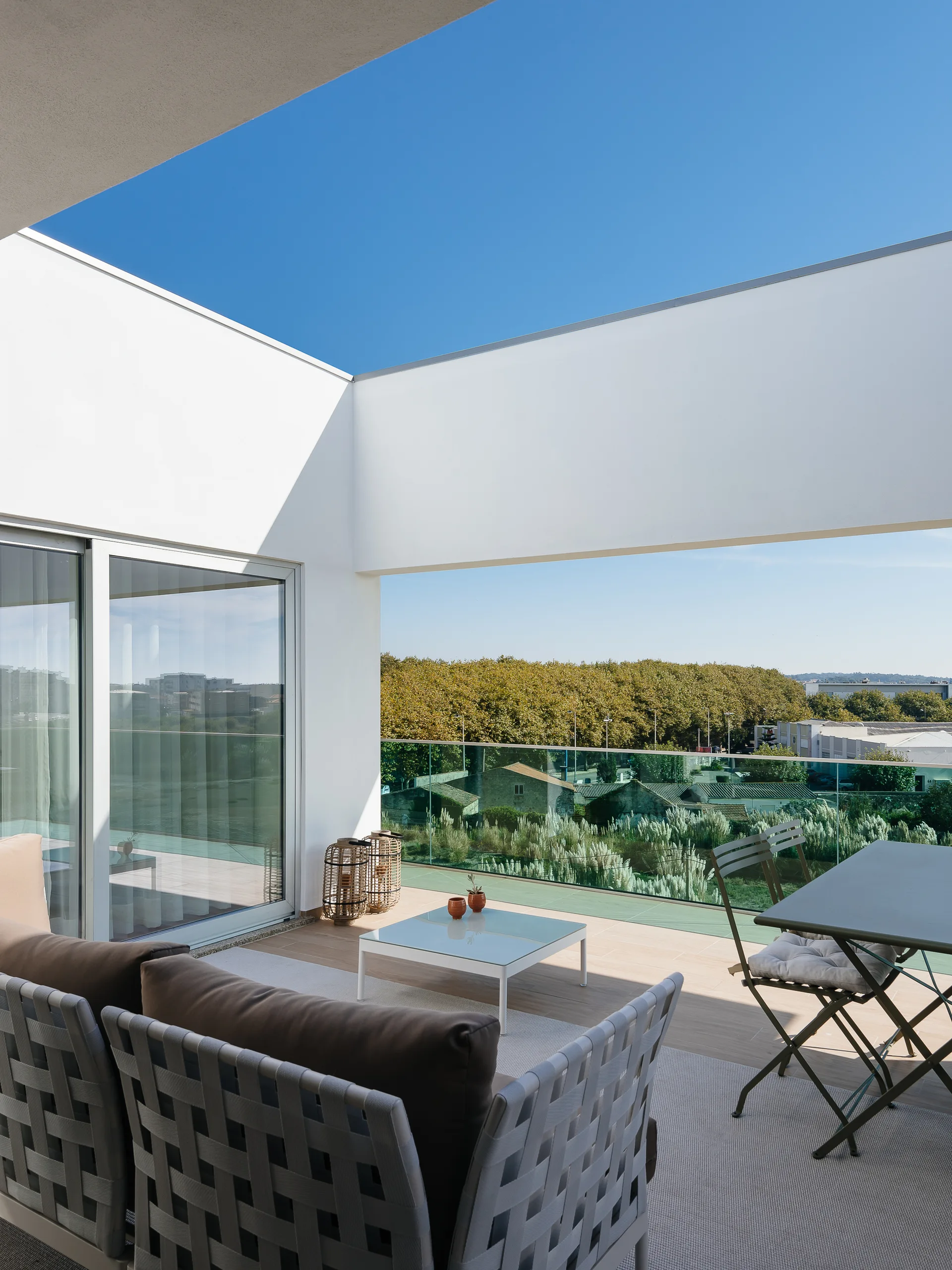 A modern balcony features a grey woven strap sofa, a white coffee table, and a glass railing overlooking a green landscape under a bright blue sky.
