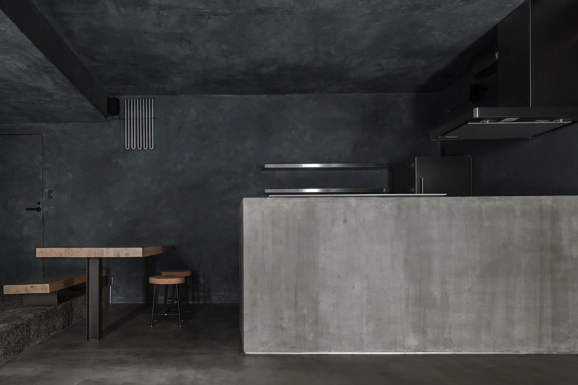 A dark minimalist kitchen features a raw concrete island, charcoal plaster walls and ceiling, and a wall-mounted wooden dining table with stools.