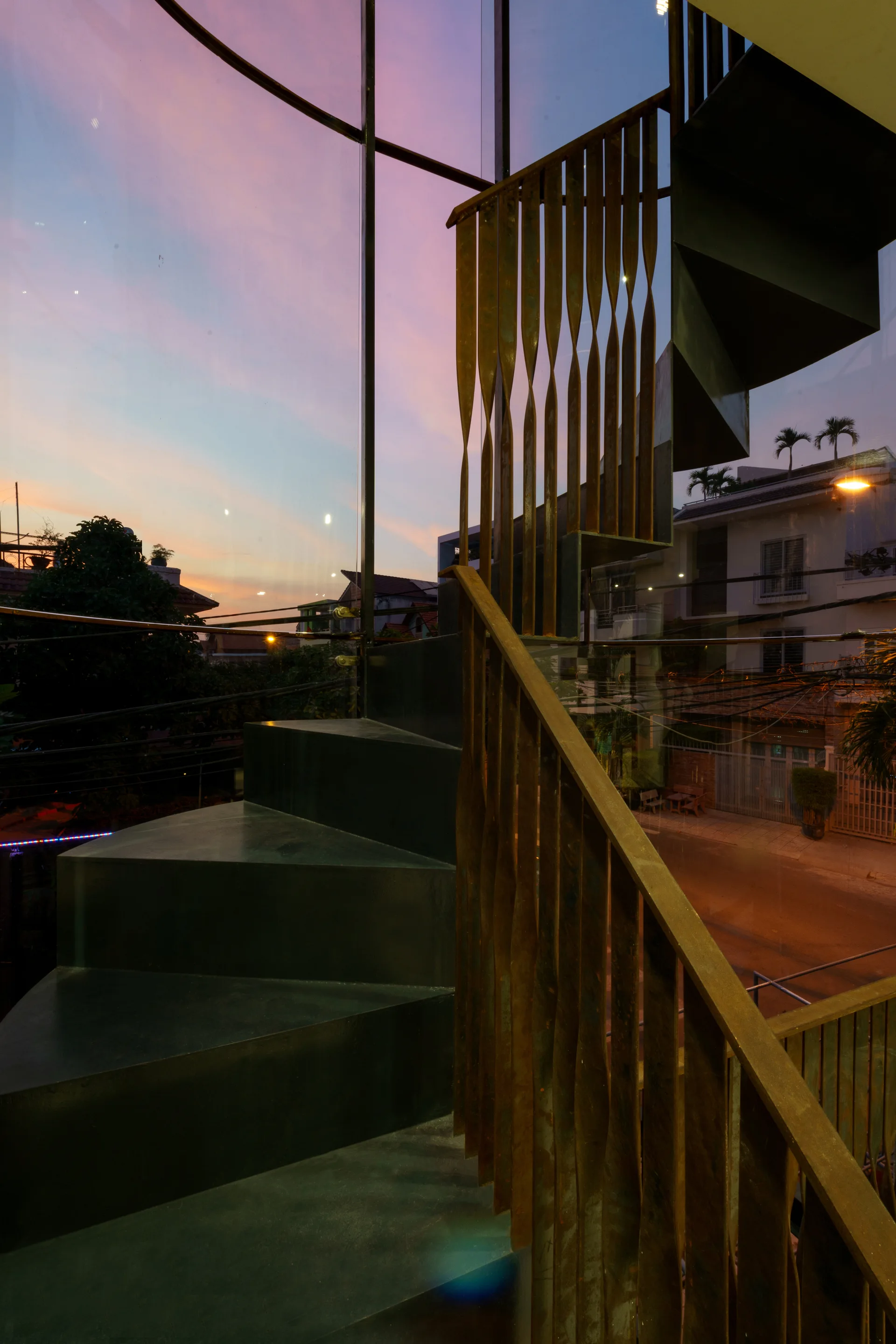 Dark metal spiral staircase with an oxidized rust-colored railing stands beside a large glass wall overlooking the street at dusk.