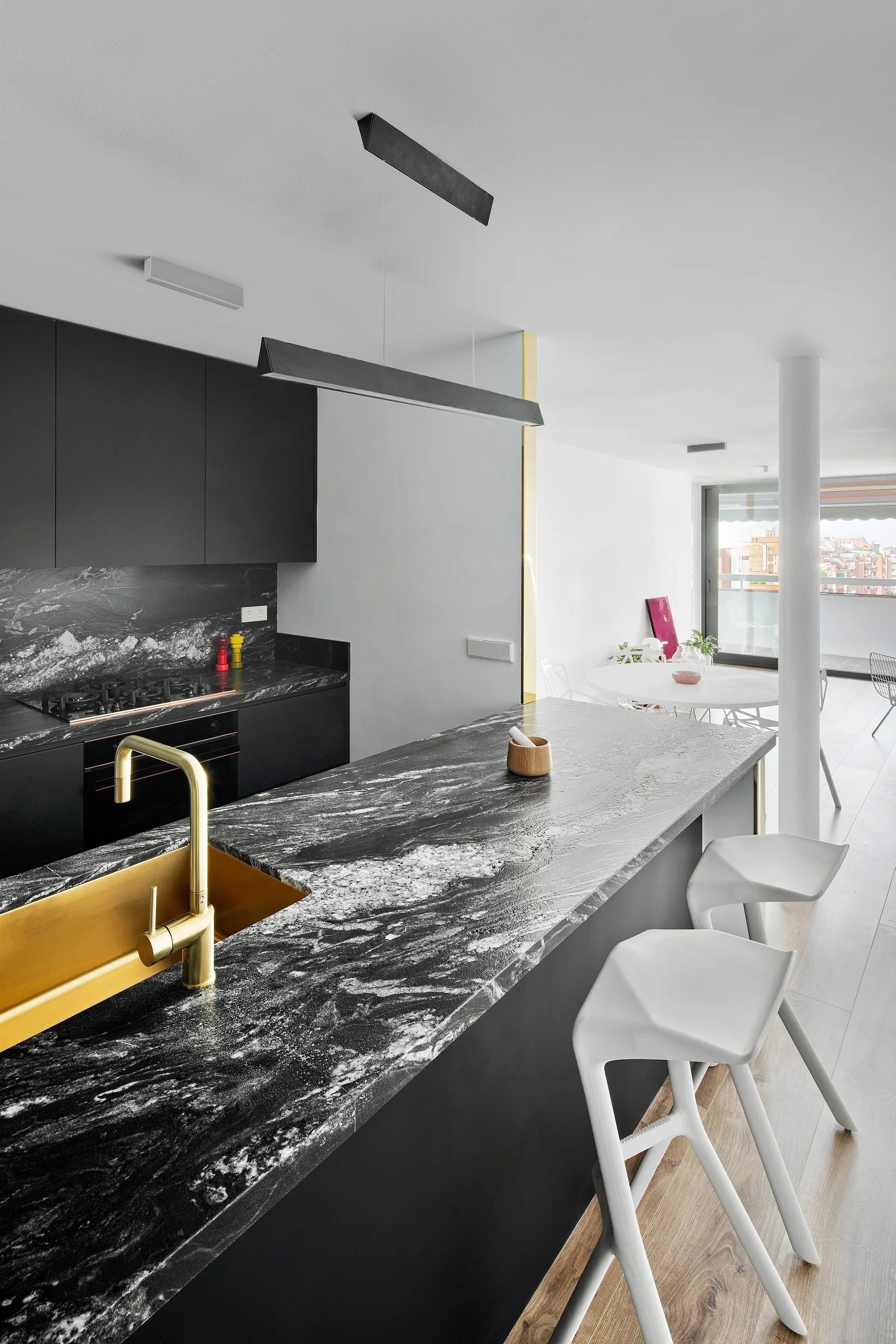 A modern kitchen features a black marble-topped island with an integrated brass sink, sculptural white bar stools, and minimalist black cabinetry.